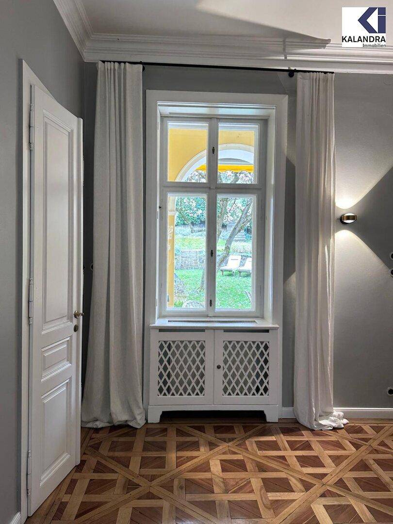 Heller Raum mit gemustertem Parkettboden, hohen Fenstern und weißer Wandverkleidung.