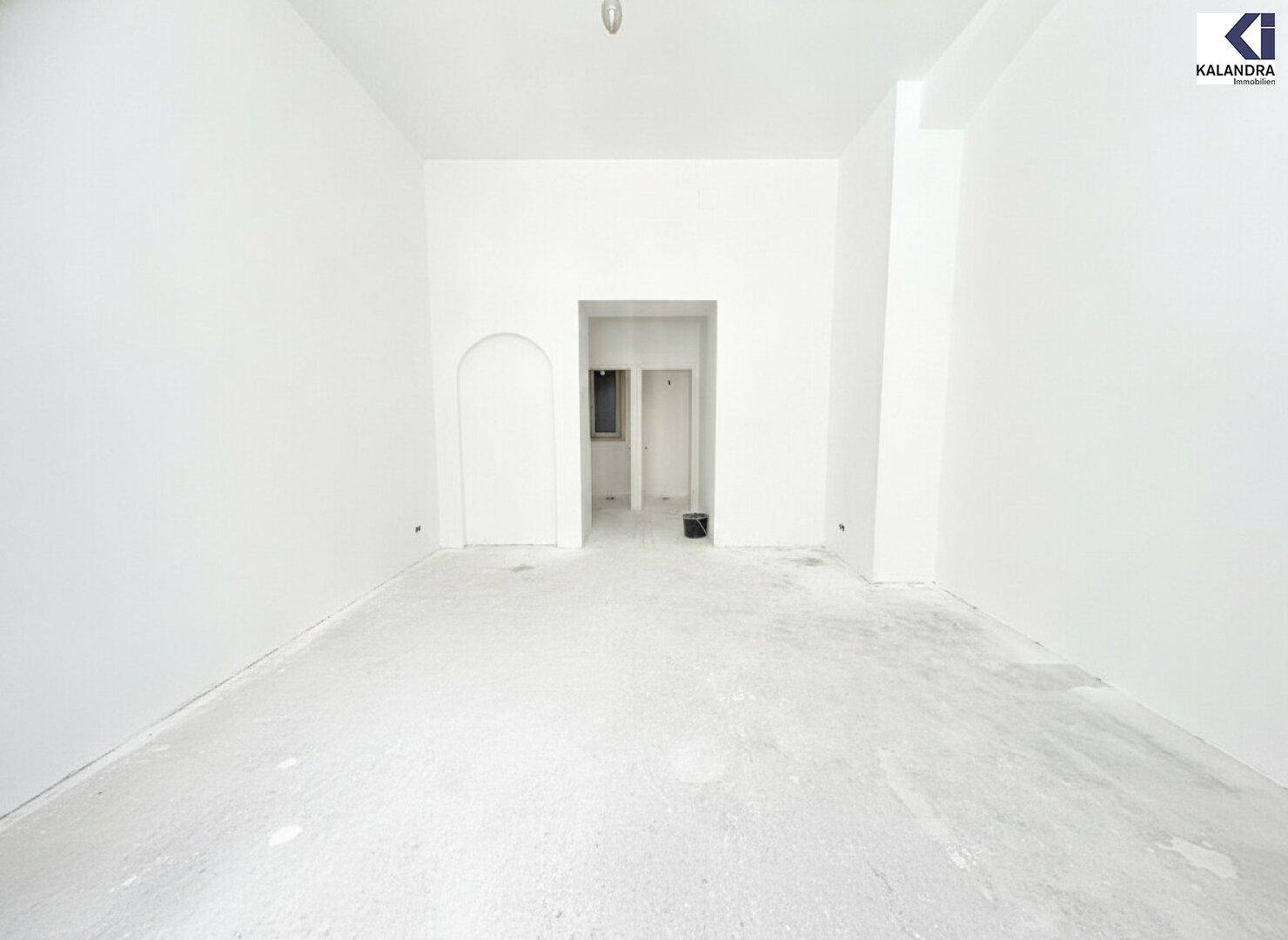 Unmöblierter Raum mit weißen Wänden, Betonboden und einem bogenförmigen Durchgang zu einem weiteren Bereich.