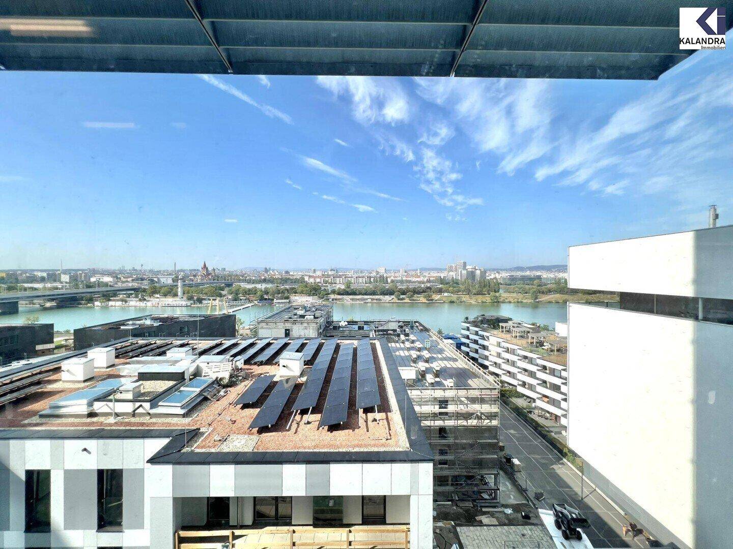 Blick aus dem Fenster auf Dächer mit Photovoltaikanlagen, einen Fluss und die Skyline der Stadt in der Ferne.