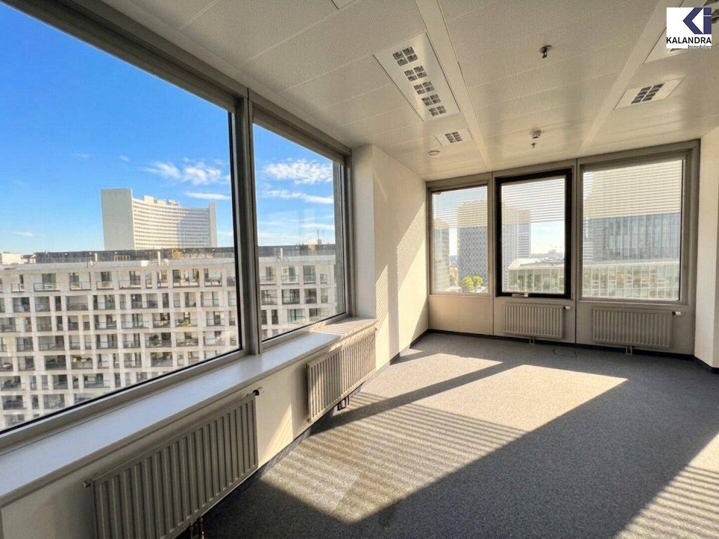 Helles, leeres Büro mit grauem Teppichboden und großen Fenstern, die einen Blick auf umliegende Gebäude ermöglichen.