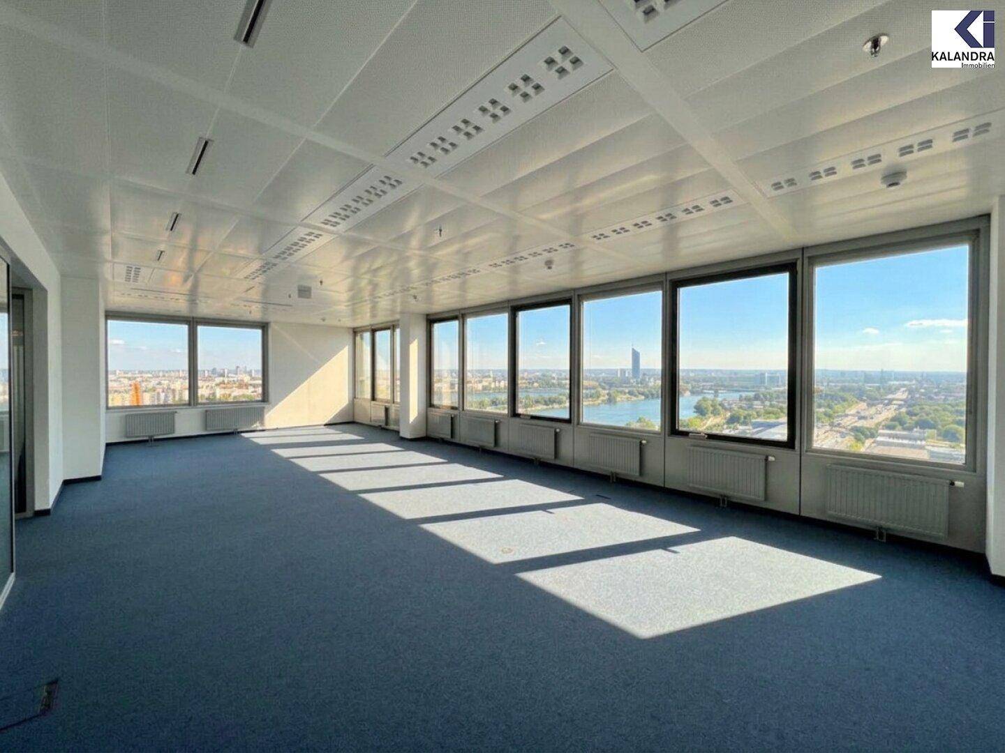 Geräumiges, leeres Büro mit blauem Teppichboden und großen Fenstern, die einen weiten Blick auf den See und die Stadt bieten.