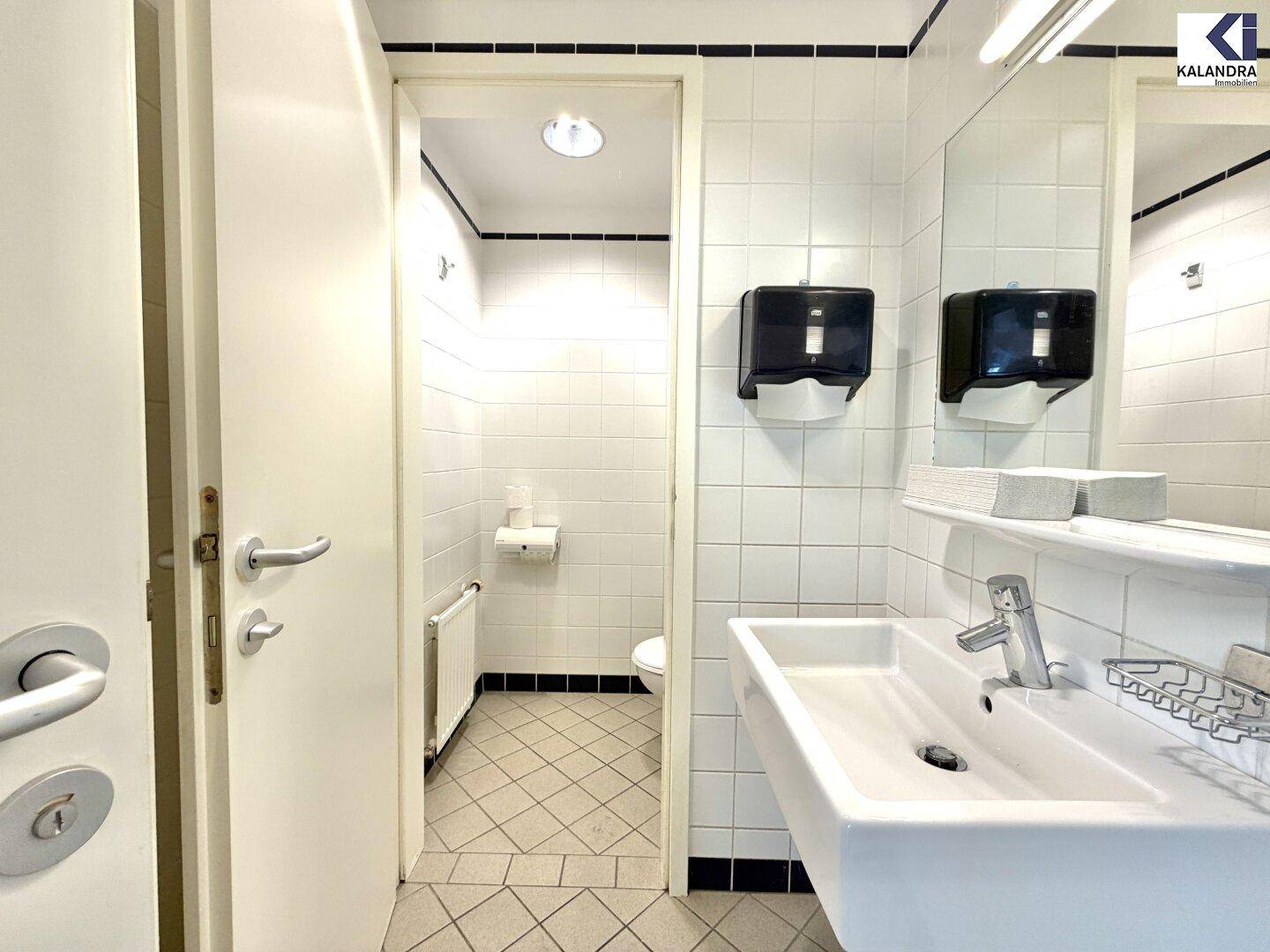 Sauberes Badezimmer mit weißen Fliesen, Waschbecken und einem separaten Bereich für die Toilette.