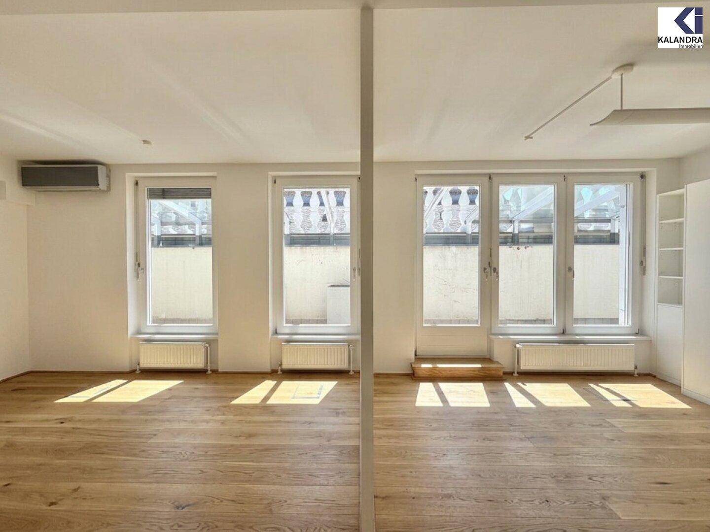 Heller Wohnraum mit Holzboden, weißen Wänden und großen Fenstern, die viel Tageslicht hereinlassen.