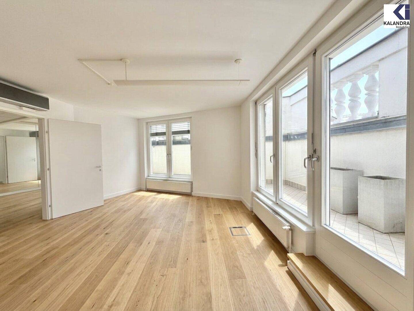 Geräumiger Raum mit Holzboden und großen Fenstern, die viel Tageslicht hereinlassen und einen Blick nach draußen ermöglichen.