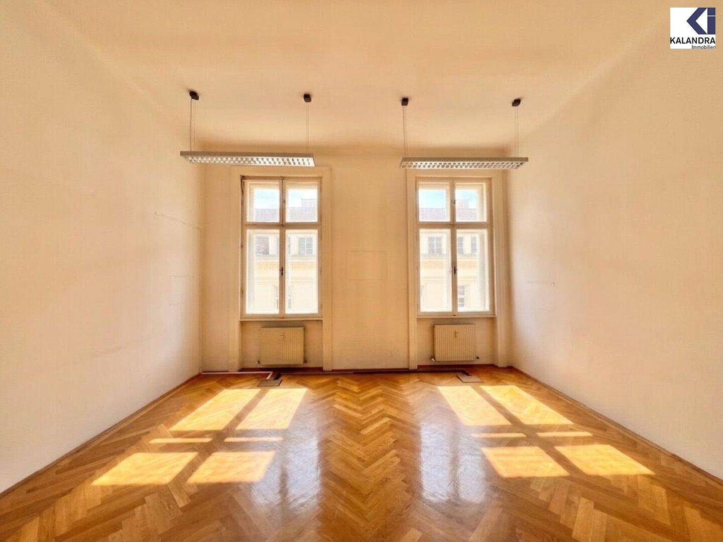 Heller Büroraum mit Fischgrätparkett und zwei großen Fenstern, die viel Licht spenden.