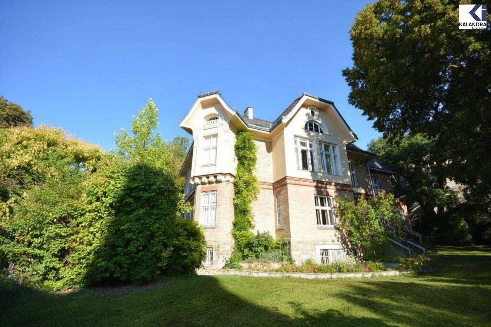 Historische Villa mit gepflegtem Garten und altem Baumbestand unter blauem Himmel.