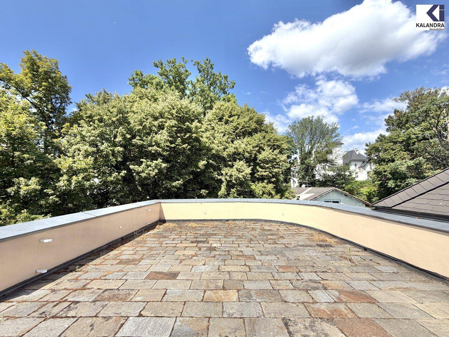 Große Dachterrasse mit Steinplattenboden und weitem Blick ins Grüne unter blauem Himmel.