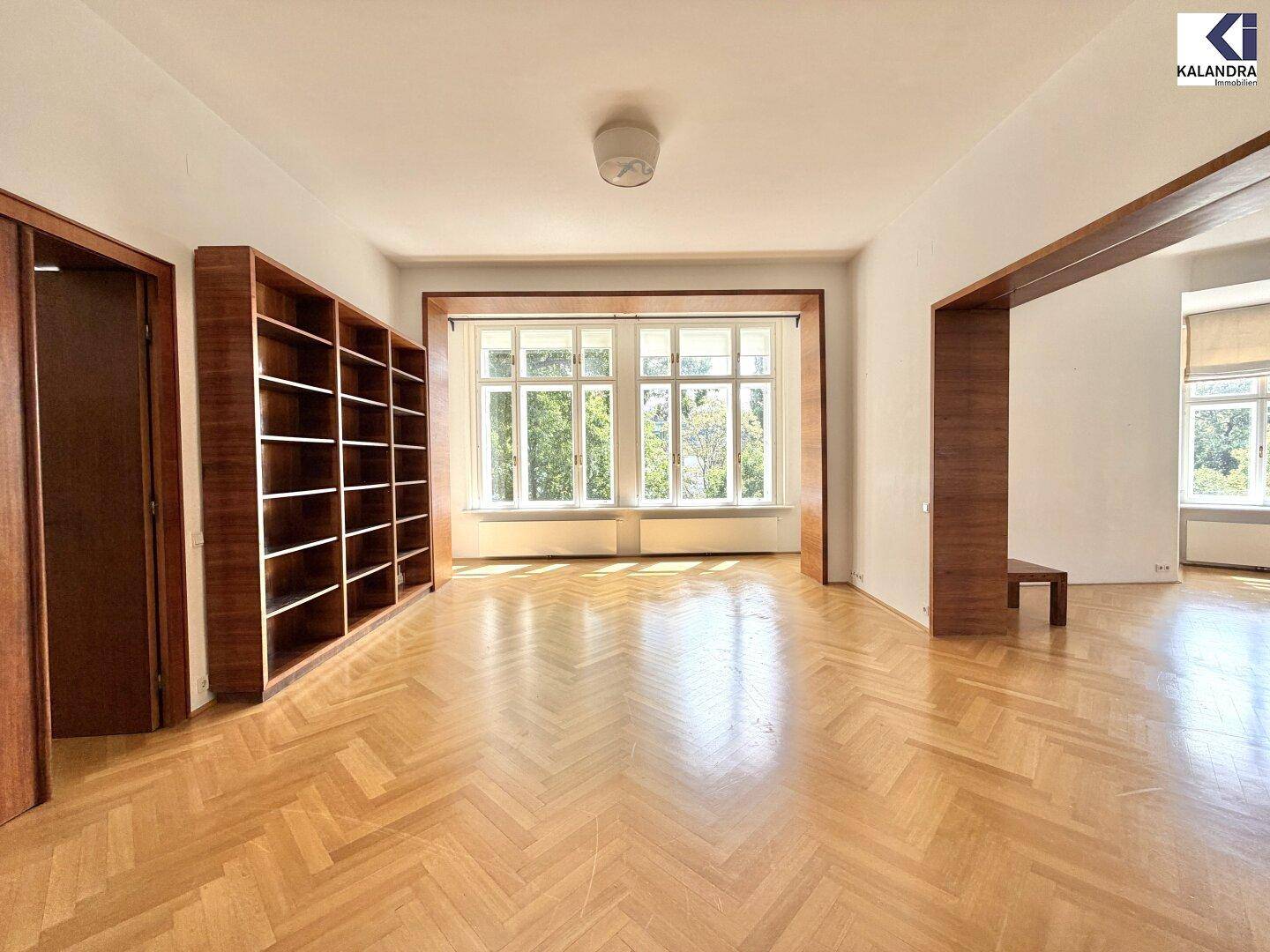 Geräumiges Wohnzimmer mit Fischgrätparkett, großen Fenstern und eingebautem Bücherregal.