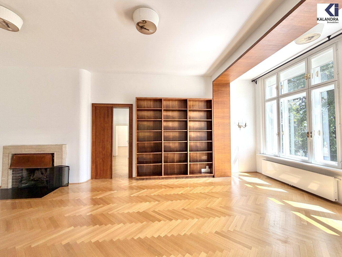 Großzügiges Wohnzimmer mit Kamin, Fischgrätparkett und Blick ins Grüne durch große Fenster.