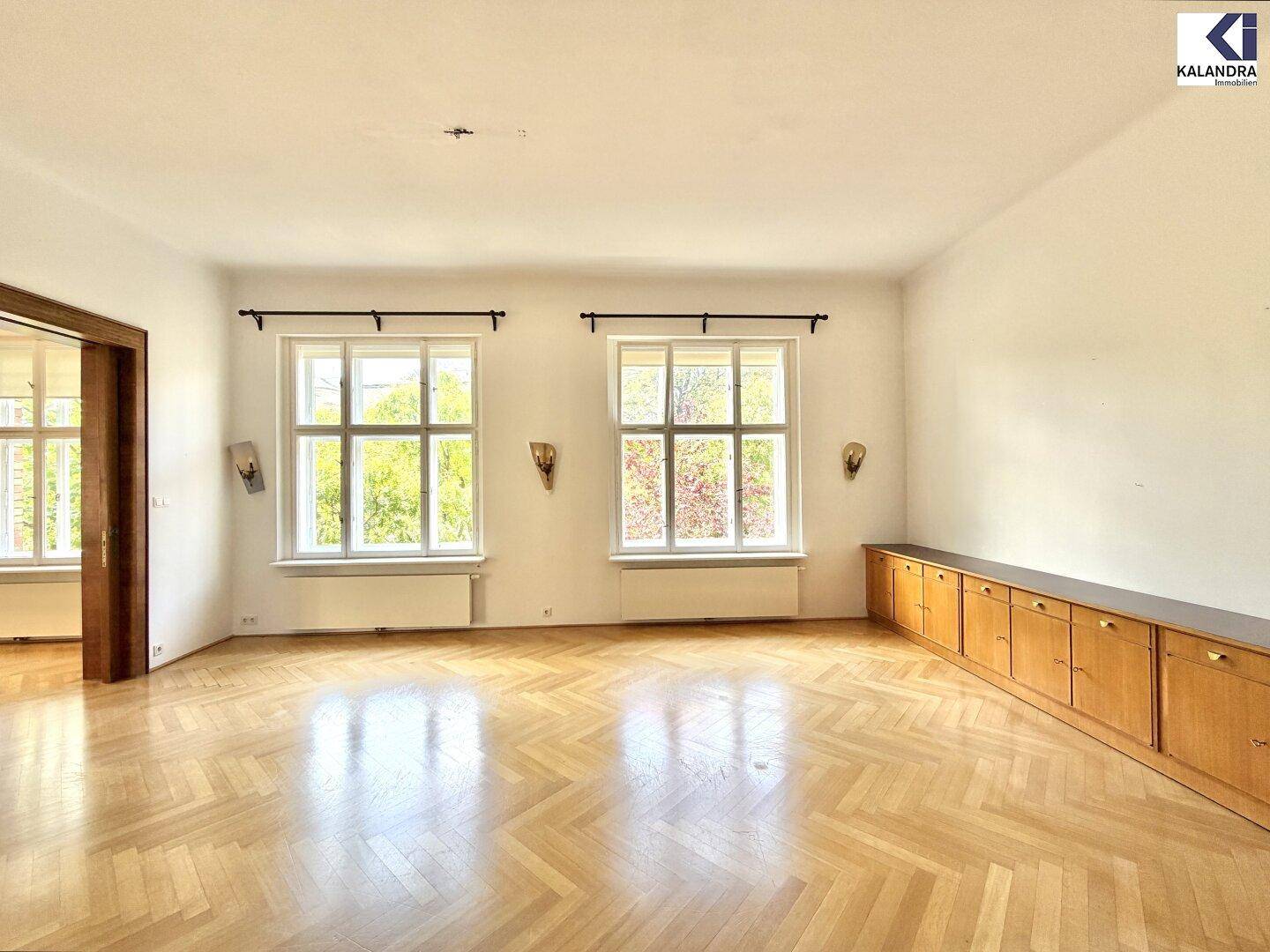 Heller Wohnbereich mit Fischgrätparkett, zwei großen Fenstern und eingebauten Sideboards.