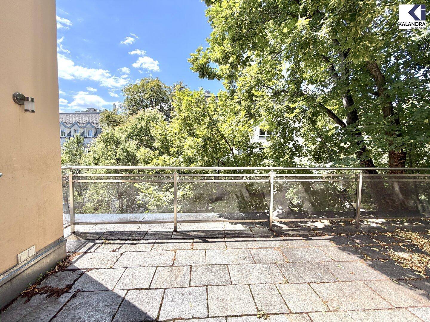 Heller Balkon mit Steinplattenboden und Glasgeländer, umgeben von hohen Bäumen.