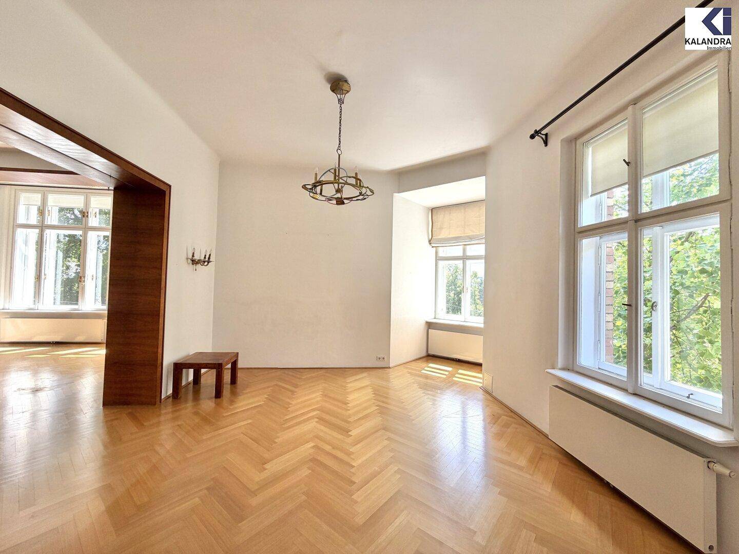 Helles Wohnzimmer mit Fischgrätparkett, Kronleuchter und großen Fenstern mit Ausblick.
