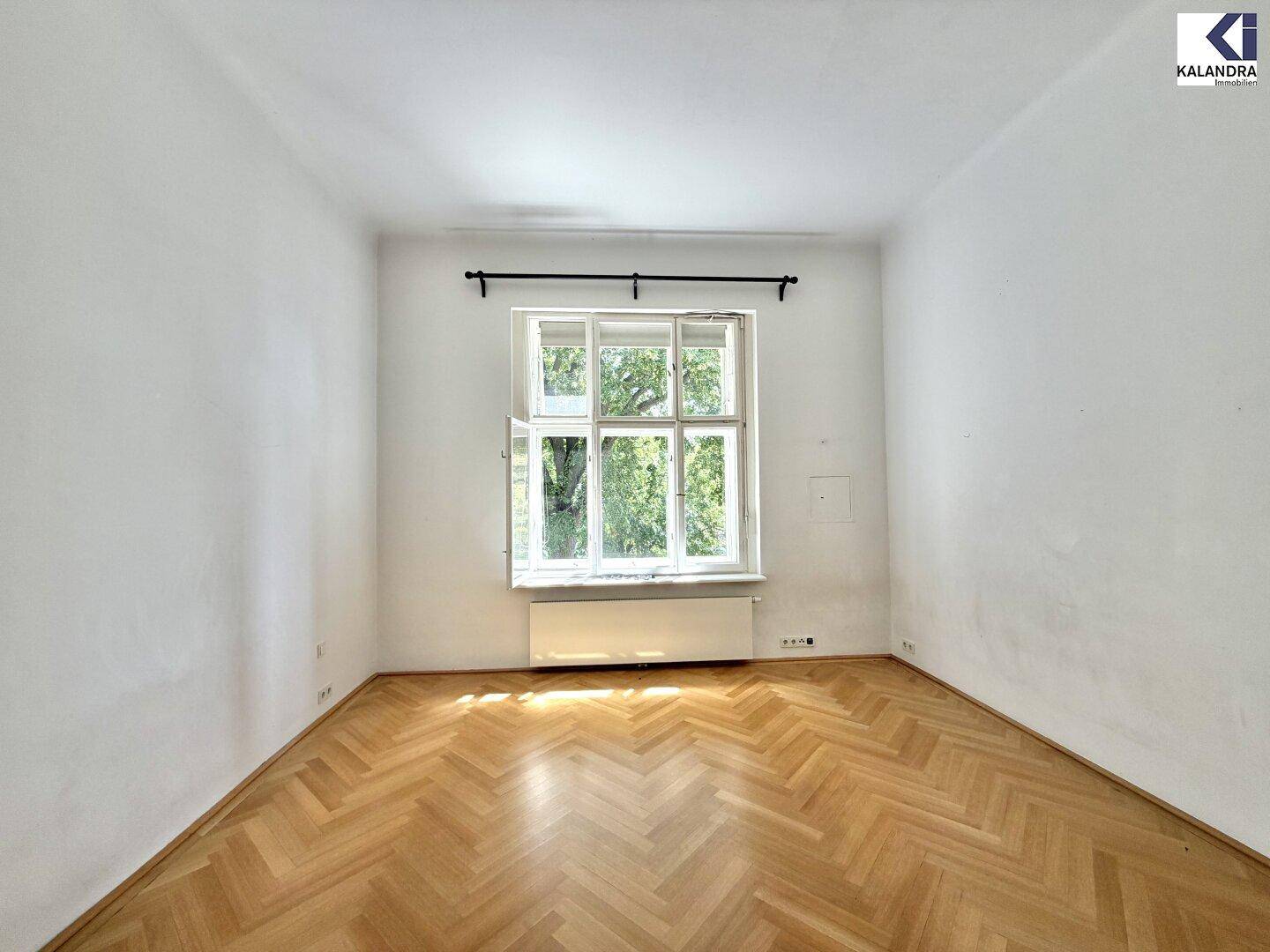 Ruhiges Zimmer mit Fischgrätparkett und einem großen Fenster mit Blick ins Grüne.