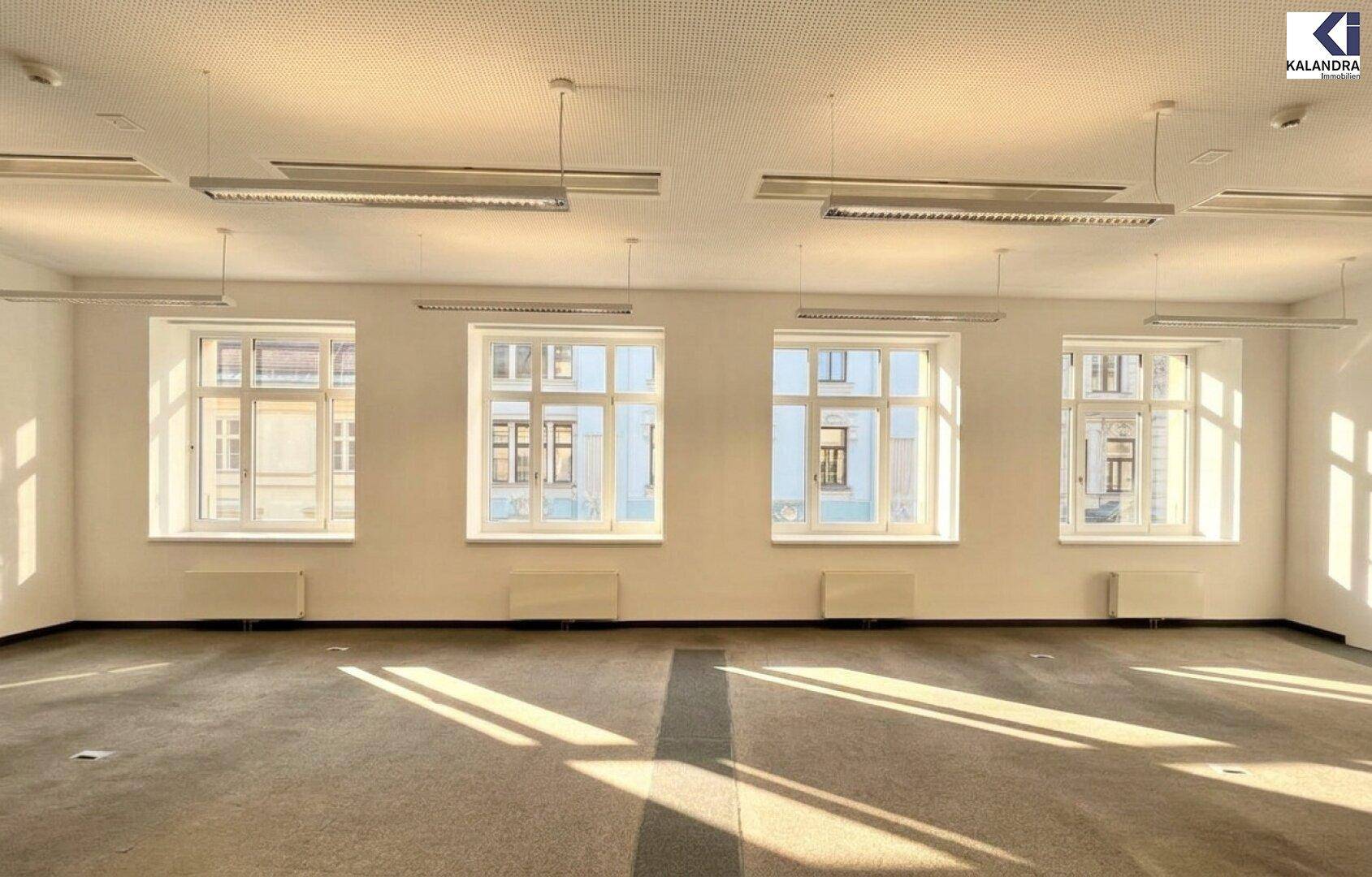 Heller Büroraum mit vier großen Fenstern, Teppichboden und viel natürlichem Tageslicht.