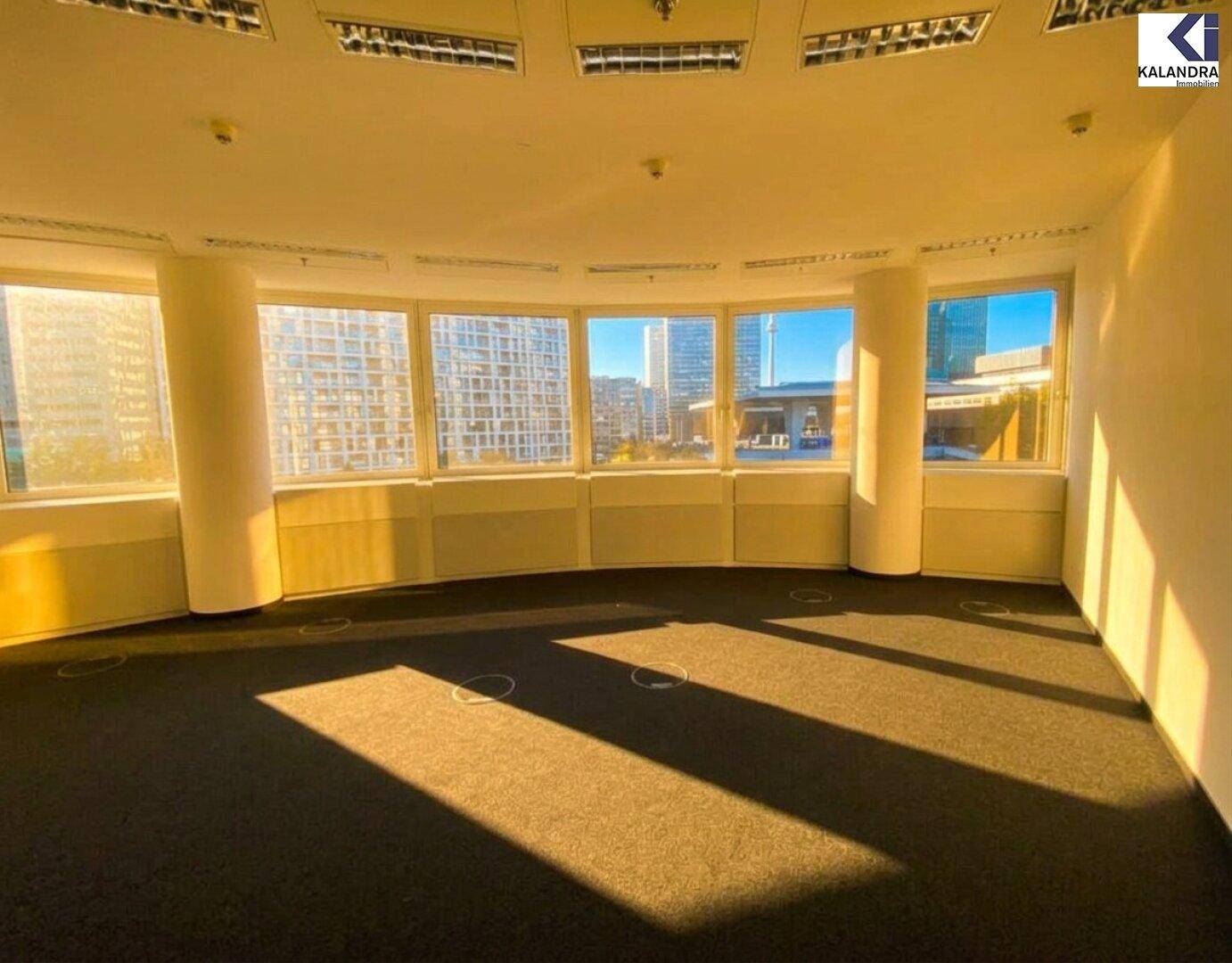 Heller Büroraum mit Teppichboden und einer Reihe großer Fenster mit Stadtpanorama, ideal für Arbeitsplätze.