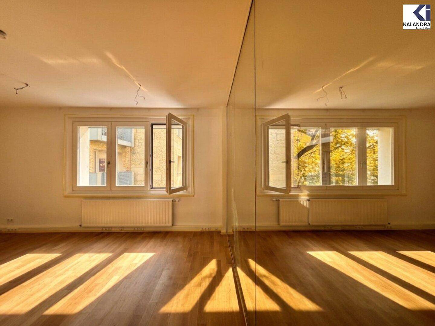 Geräumiger Wohnbereich mit Holzboden, großen Fenstern und einer spiegelnden Wand, die den Raum erweitert.