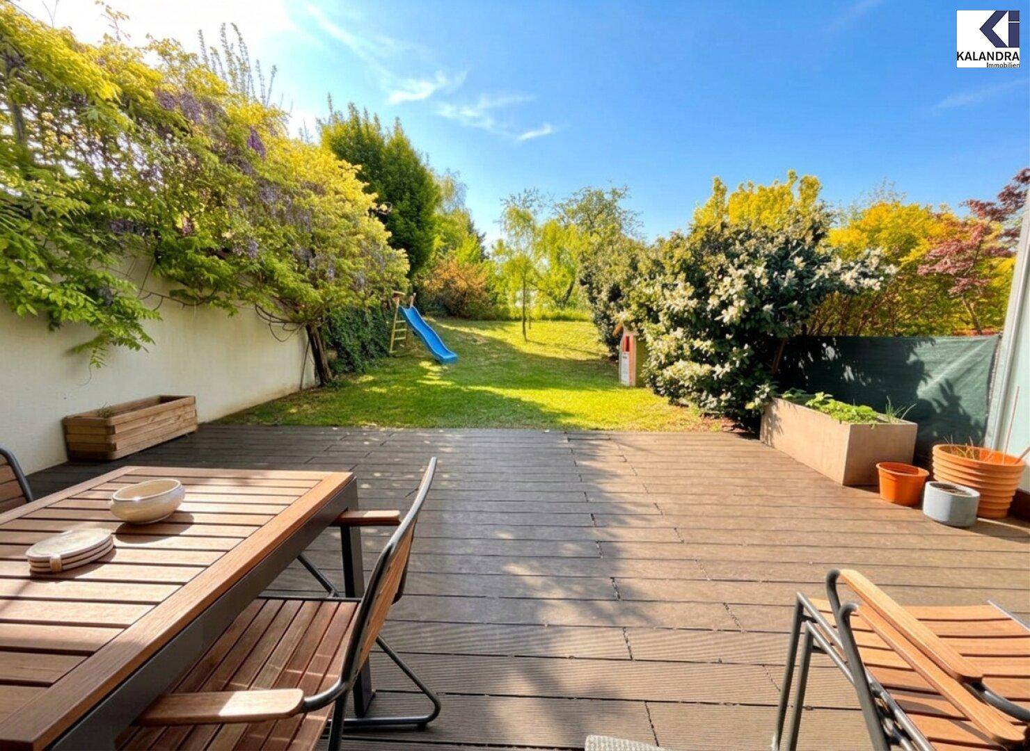 Sonnige Holzterrasse mit Gartenblick und kleiner Kinderrutsche im Hintergrund.