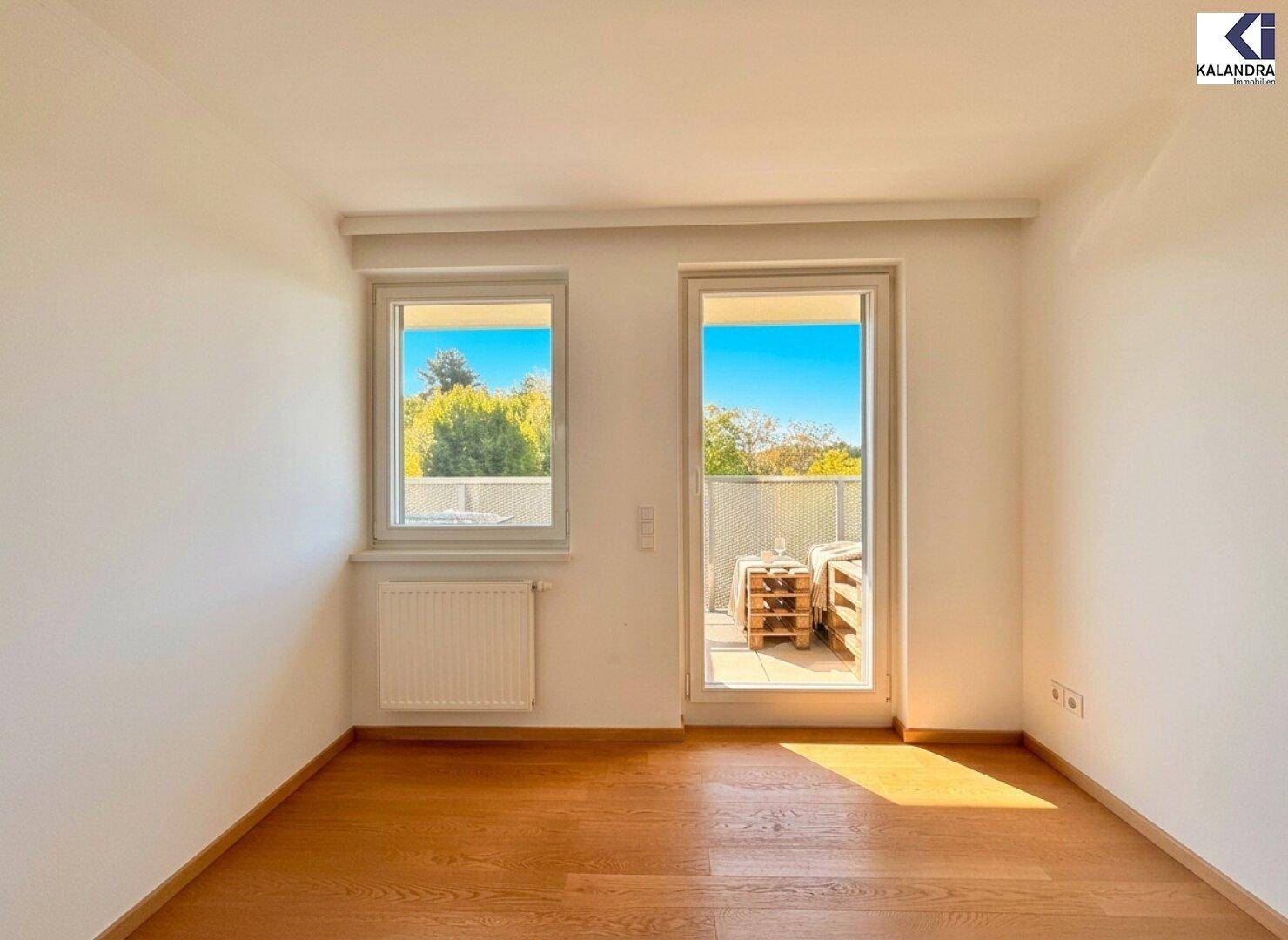Leeres Zimmer mit Holzboden und Zugang zu einem sonnigen Balkon.