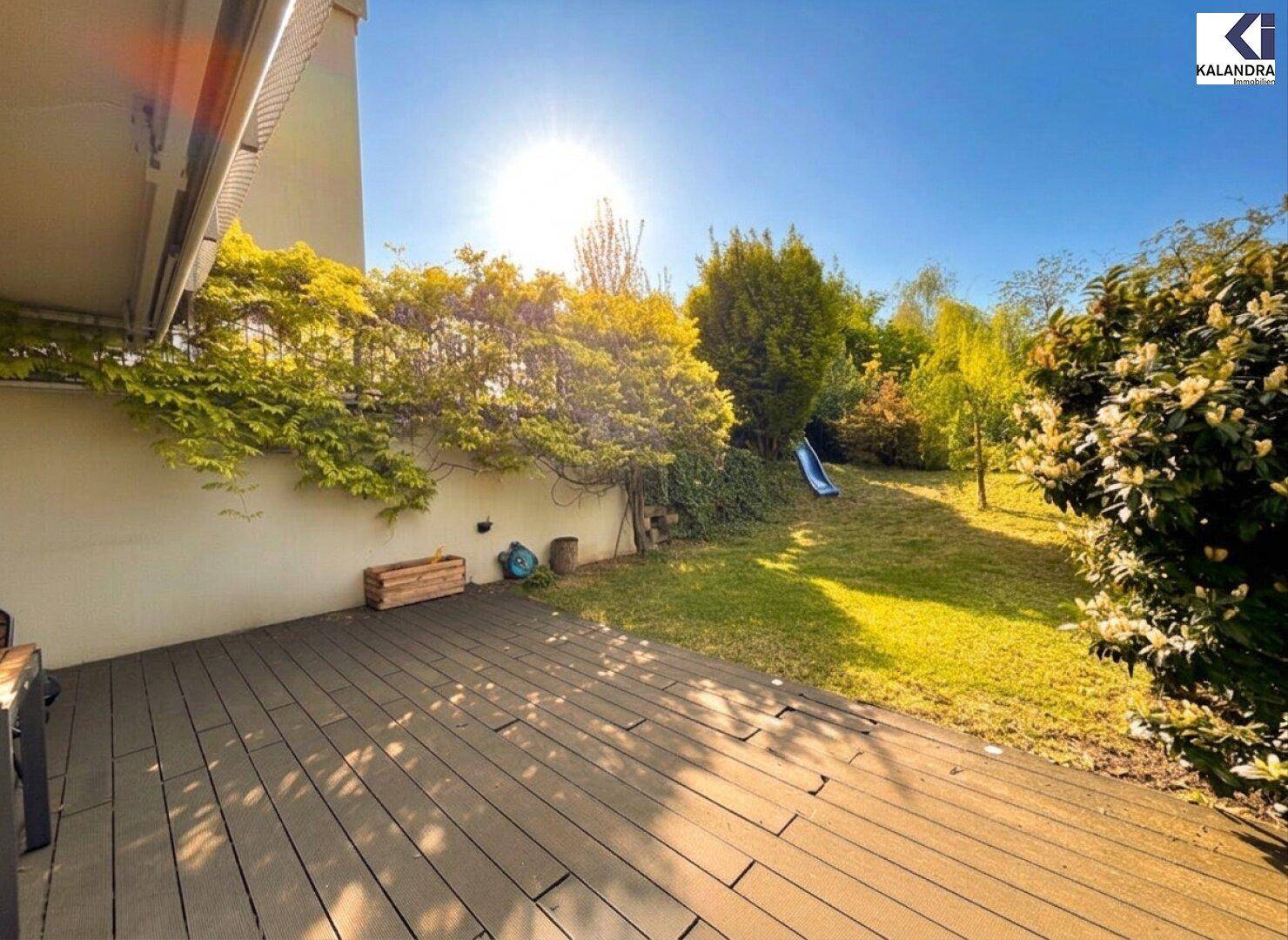Sonnige Holzterrasse mit Zugang zum grünen Garten und blühenden Pflanzen.