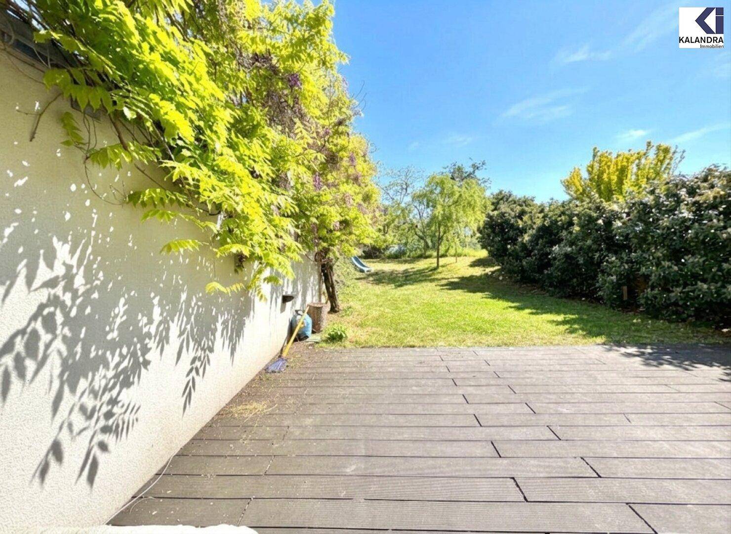 Sonnige Terrasse mit Holzbelag und Blick auf den grünen Garten mit Bäumen und Spielbereich.