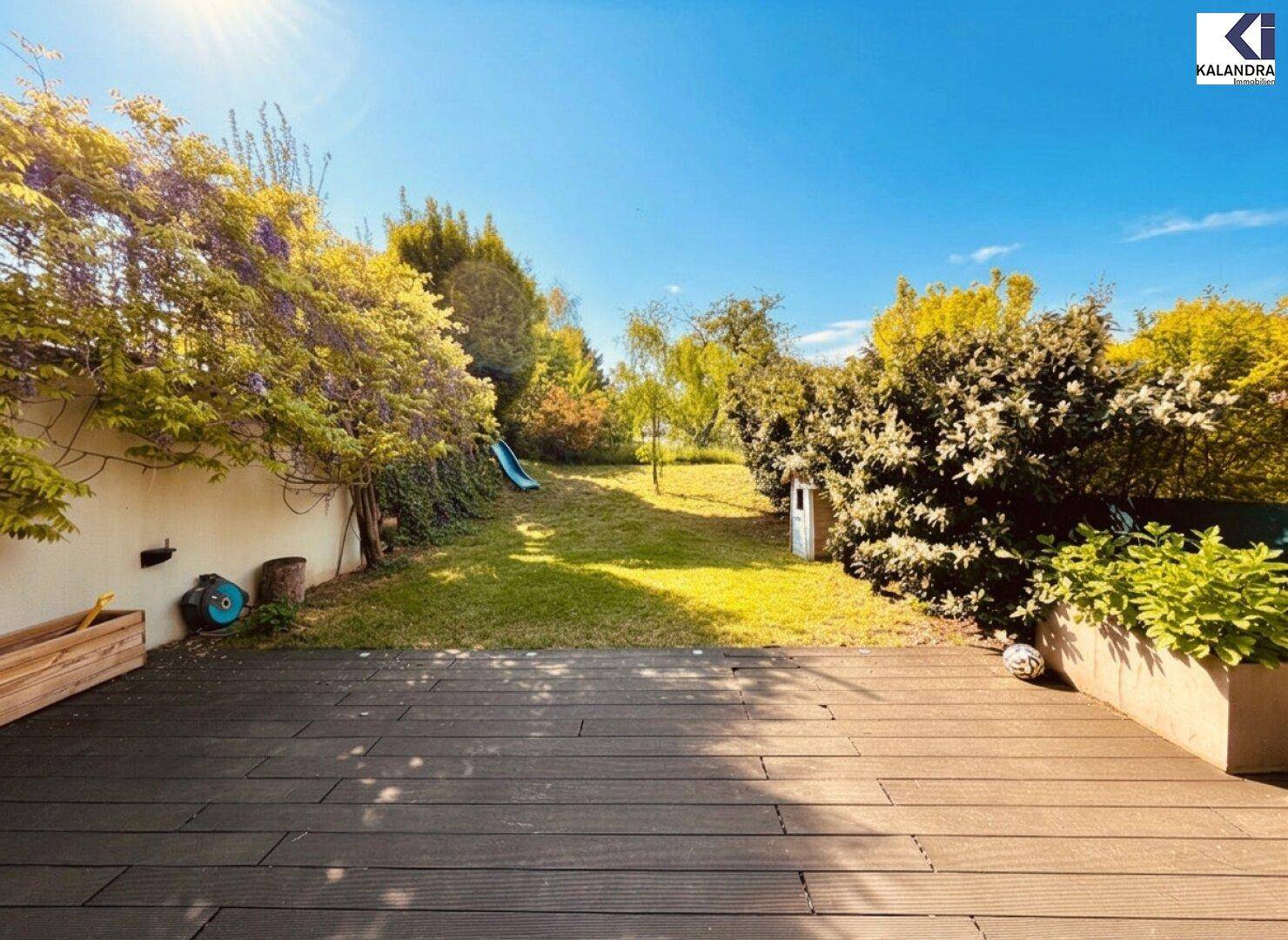 Holzterrasse mit Blick auf den grünen Garten und eine kleine Rutsche im Hintergrund.