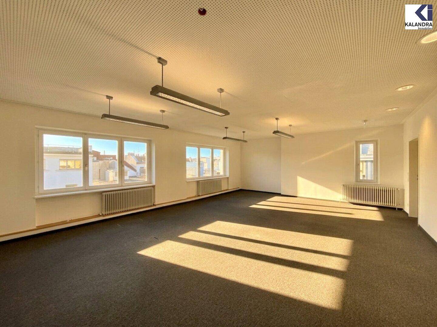 Geräumiger Bürobereich mit Teppichboden, großen Fenstern und modernen Deckenleuchten, ideal für Arbeitsplätze.