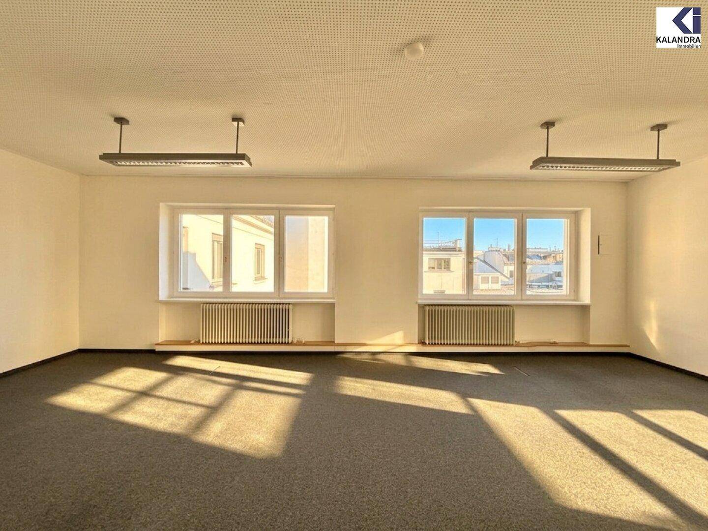 Heller Büroraum mit Teppichboden und zwei großen Fenstern, die viel Tageslicht hereinlassen.