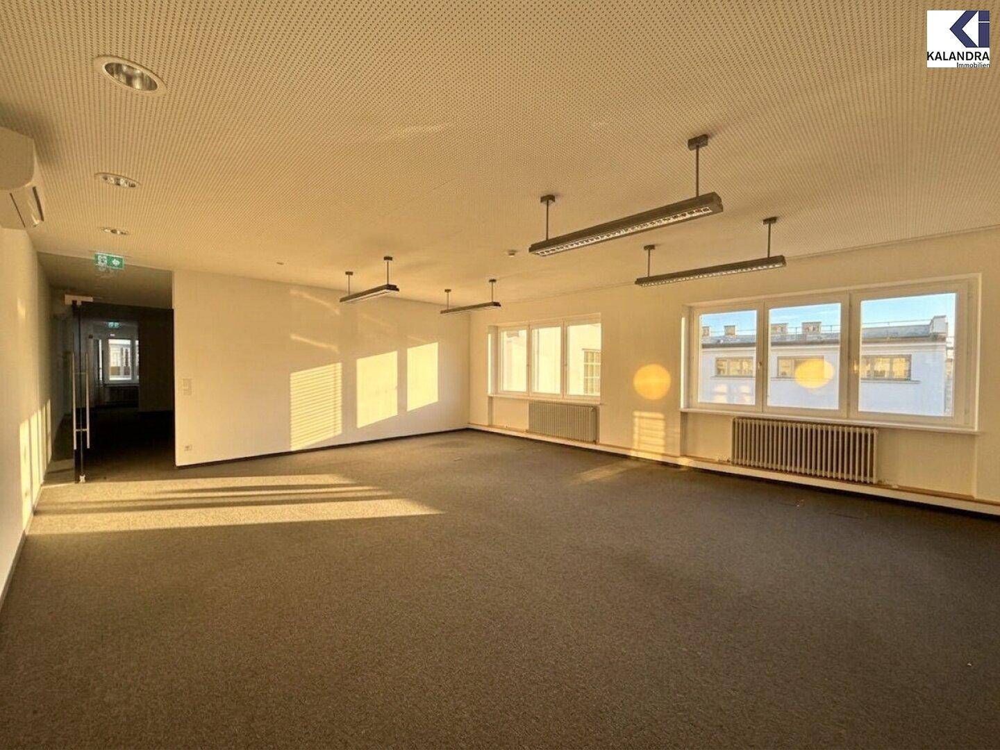 Großer, heller Büroraum mit Teppichboden und mehreren Fenstern, die für eine angenehme Arbeitsatmosphäre sorgen.