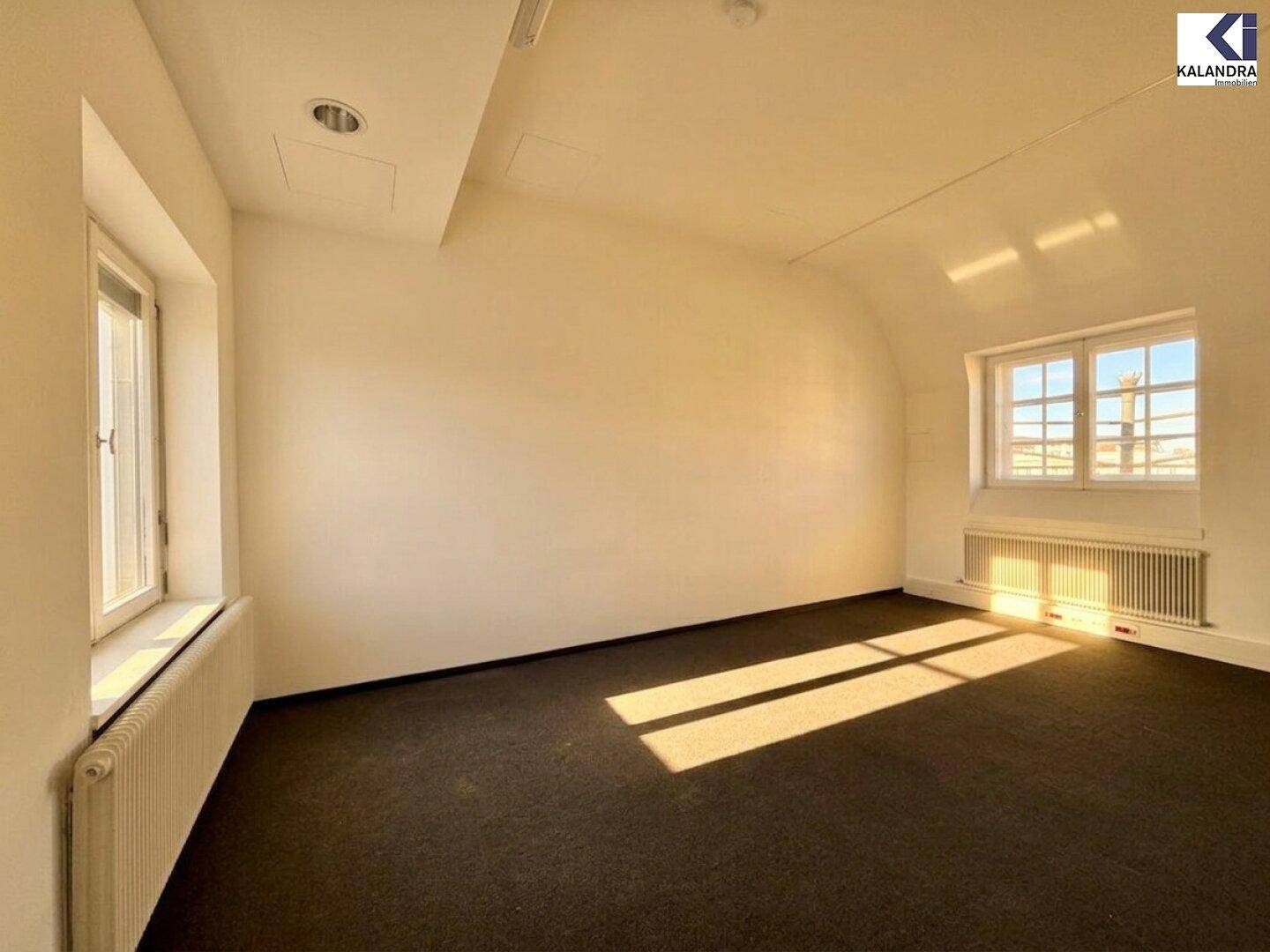 Kleinerer Büroraum mit Teppichboden, schrägen Wänden und Fenstern, die viel Sonnenlicht hereinlassen.