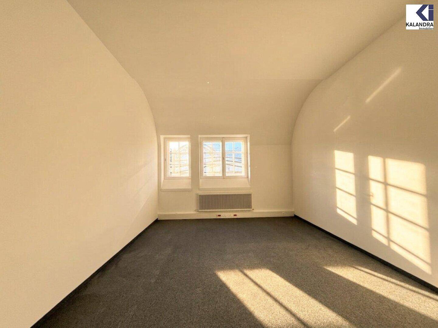 Kleinerer Büroraum mit Teppichboden, schrägen Wänden und zwei Fenstern, die viel Sonnenlicht hereinlassen.