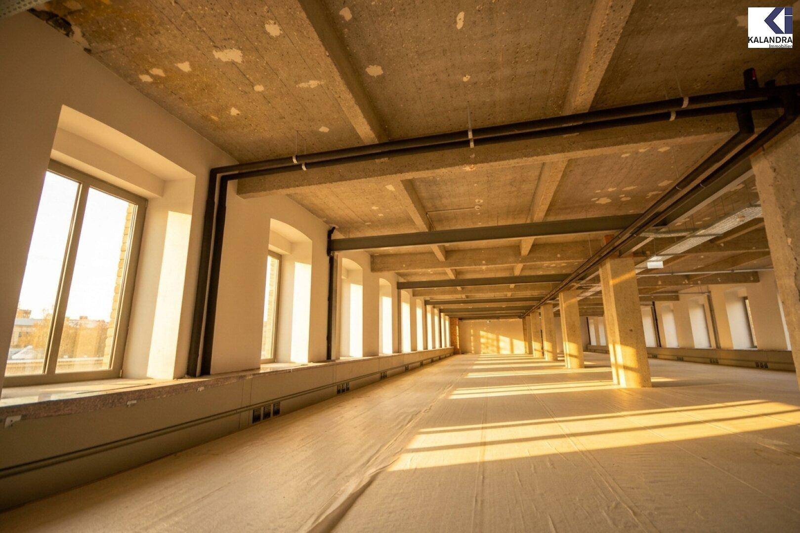 Langer, offener Büroraum mit vielen Fenstern entlang der Wand und sichtbaren Deckenstrukturen.