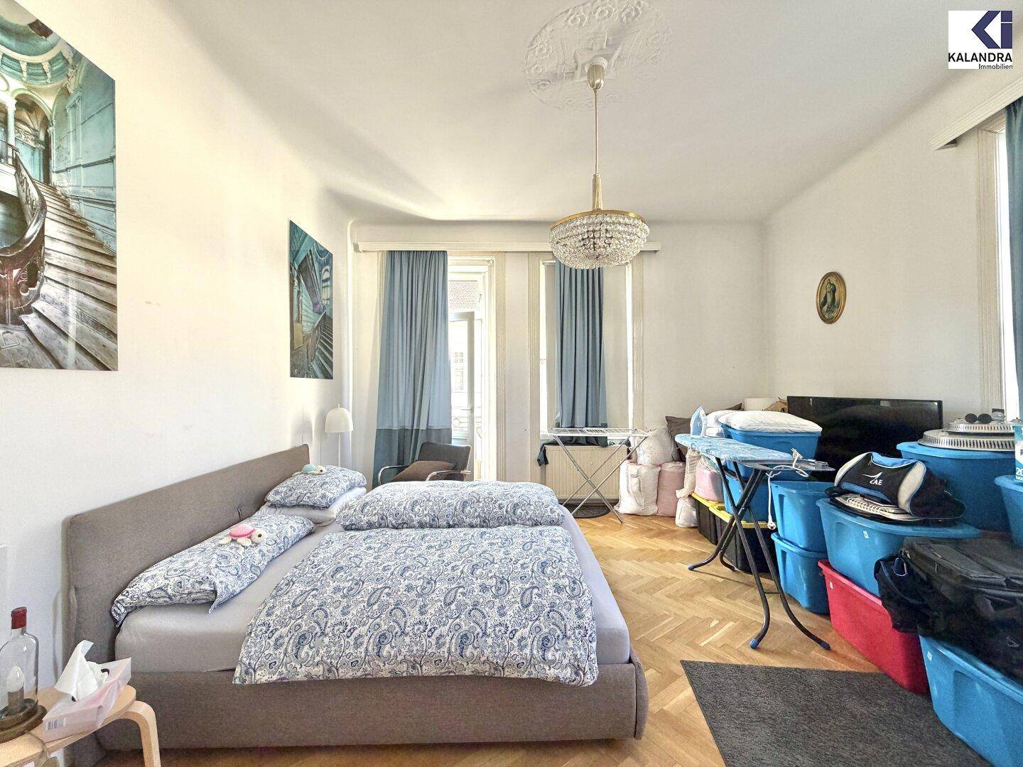 Schlafzimmer mit Fischgrätparkett, Kronleuchter und Blick auf die Straße durch große Fenster.