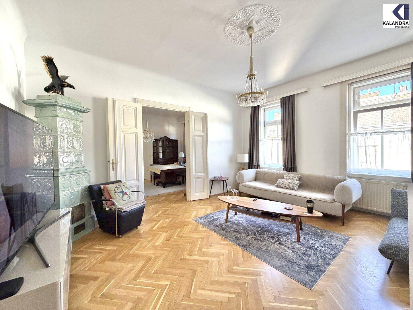 Geräumiges Wohnzimmer mit traditionellem Kachelofen, Fischgrätparkett und Blick in den Essbereich.