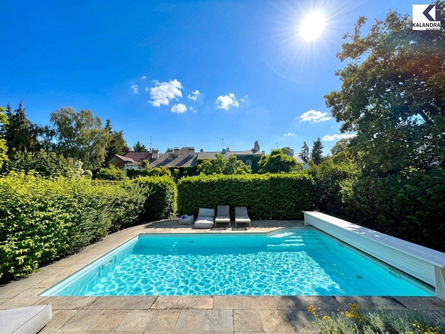 Sonnige Poolanlage mit Liegestühlen und gepflegter Hecke, umgeben von üppigem Grün und Häusern im Hintergrund.