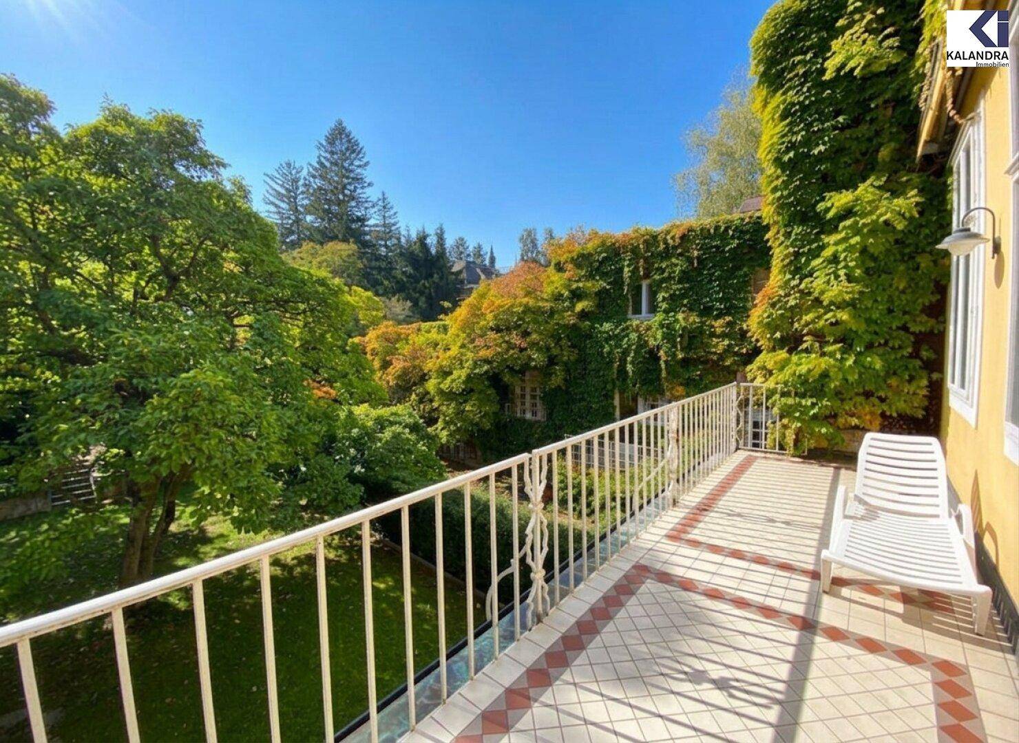 Heller Balkon mit weißem Geländer und weitem Blick auf den grünen Garten und die Rückseite der Villa.