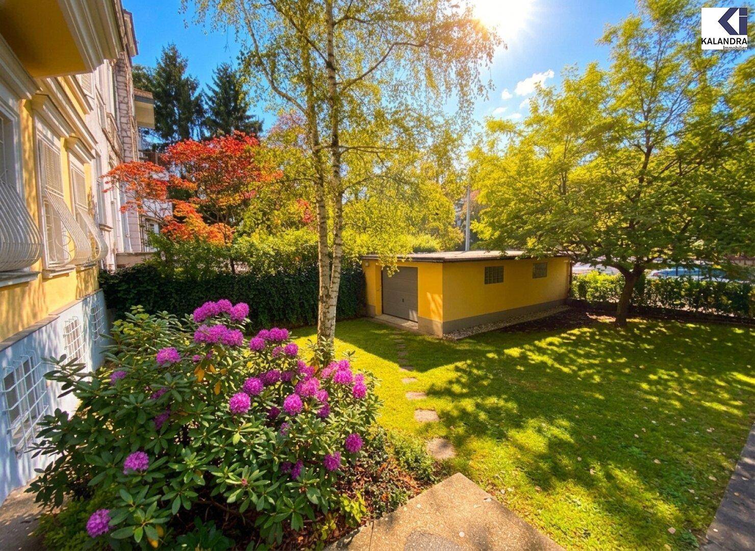 Gepflegter Vorgarten mit blühendem Rhododendron, einem kleinen Schuppen und alten Bäumen im Sonnenlicht.