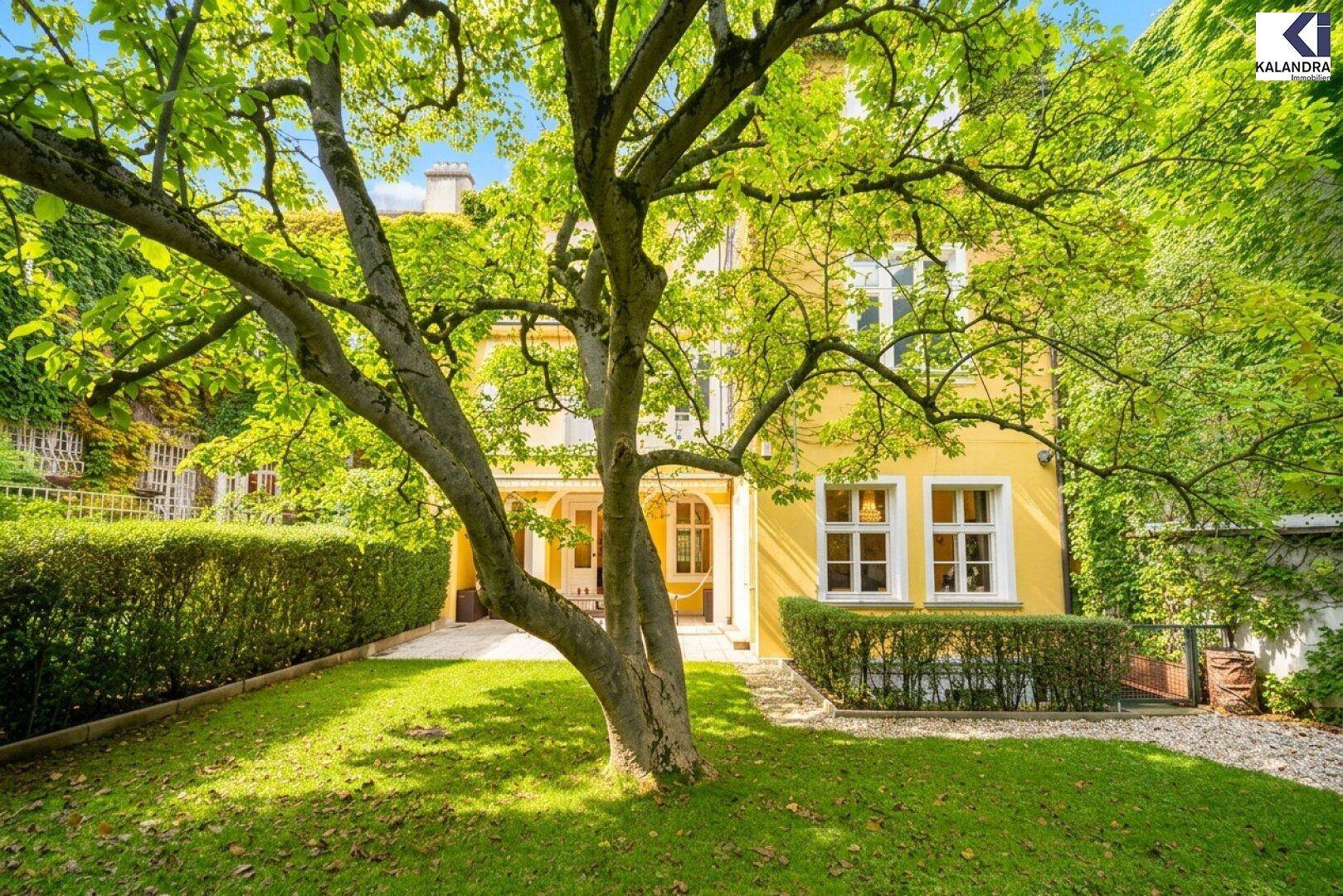 Großer, gepflegter Garten mit einem imposanten alten Baum und dichten Hecken vor der gelben Villa.