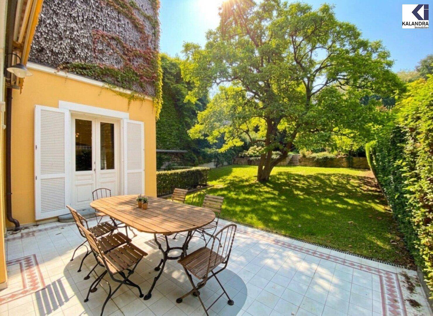 Sonniger Essbereich im Freien auf einer gefliesten Terrasse mit direktem Zugang zum Haus und Blick auf den Garten.