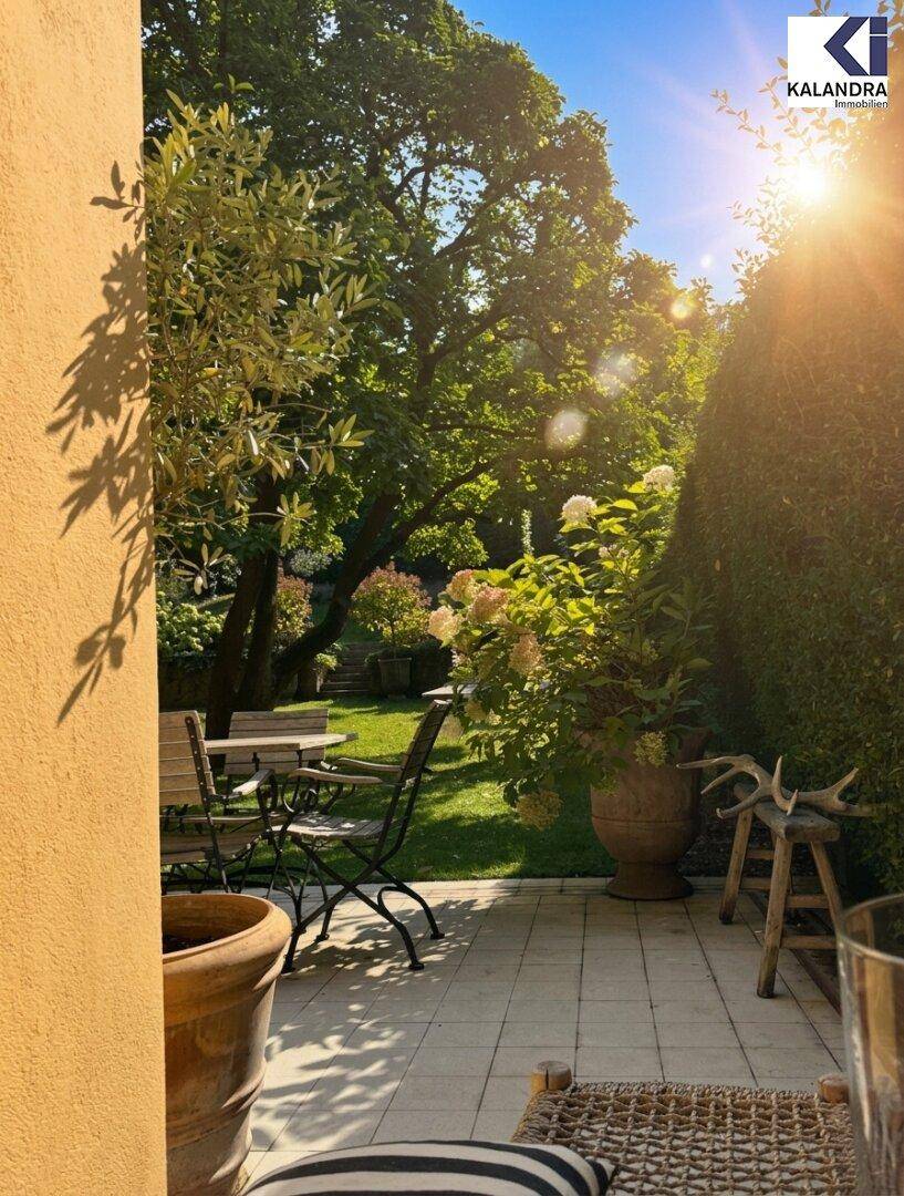 Gemütlicher Außenbereich mit Tisch und Stühlen, umgeben von üppigem Grün und alten Bäumen im Sonnenlicht.