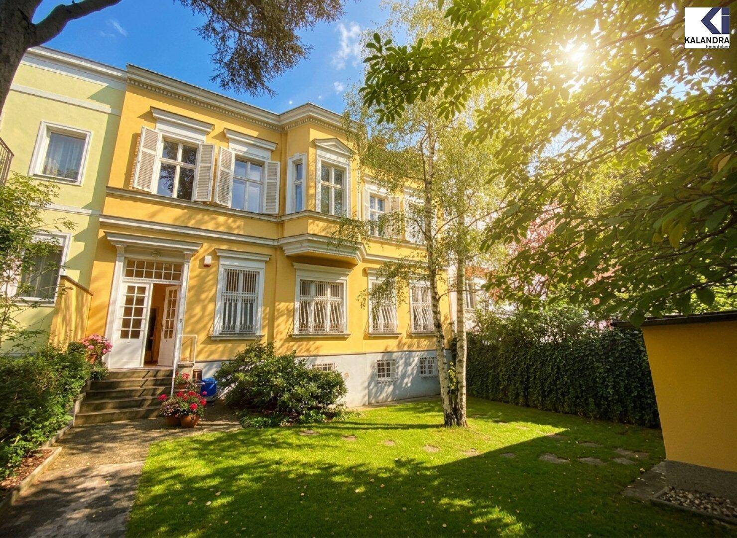 Historische gelbe Villa mit weißen Fensterläden und einem gepflegten Vorgarten unter strahlendem Sonnenschein.