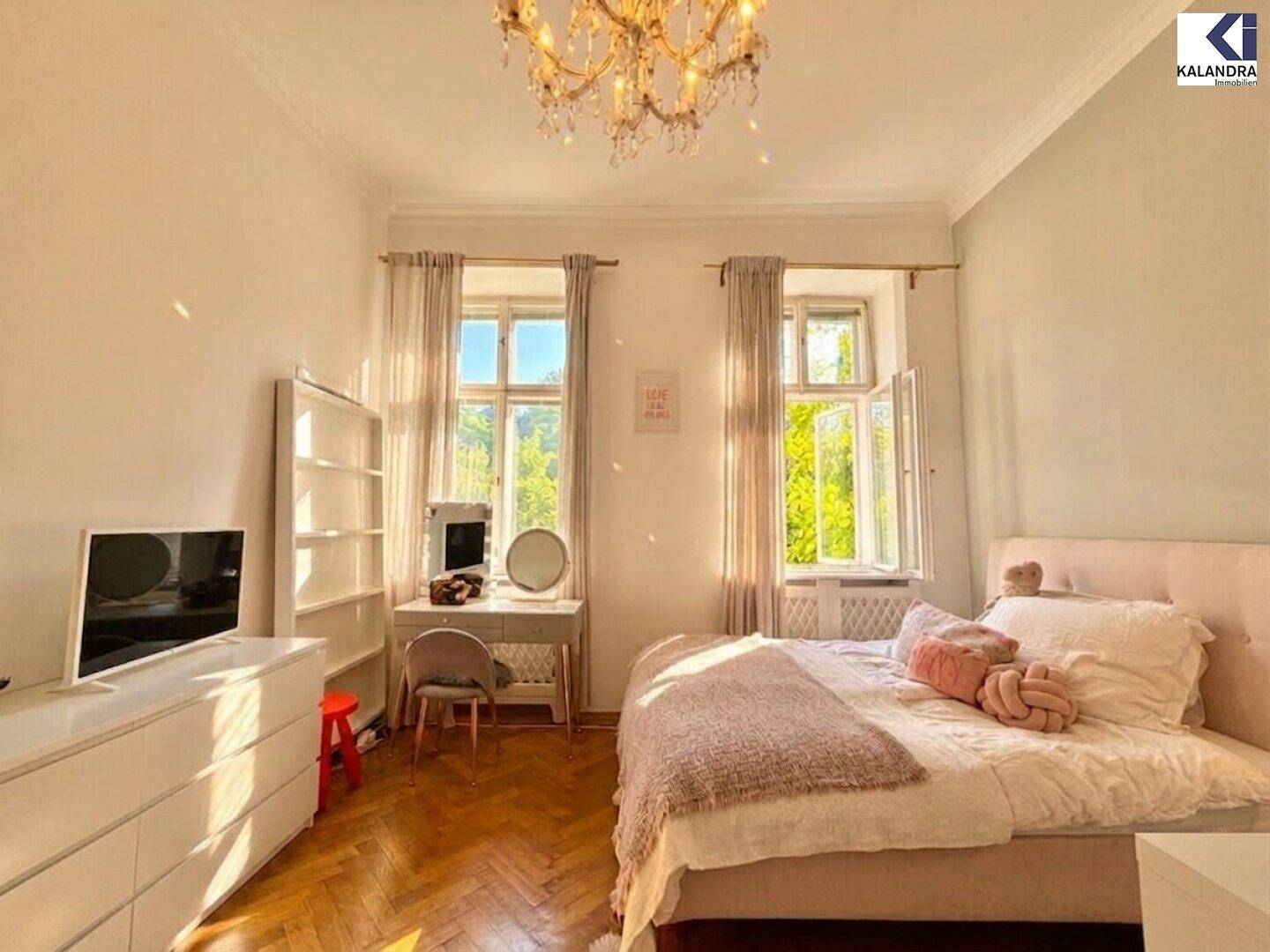 Helles Schlafzimmer mit Bett, Kommode, Schminktisch und Parkettboden, durchflutet von Sonnenlicht.