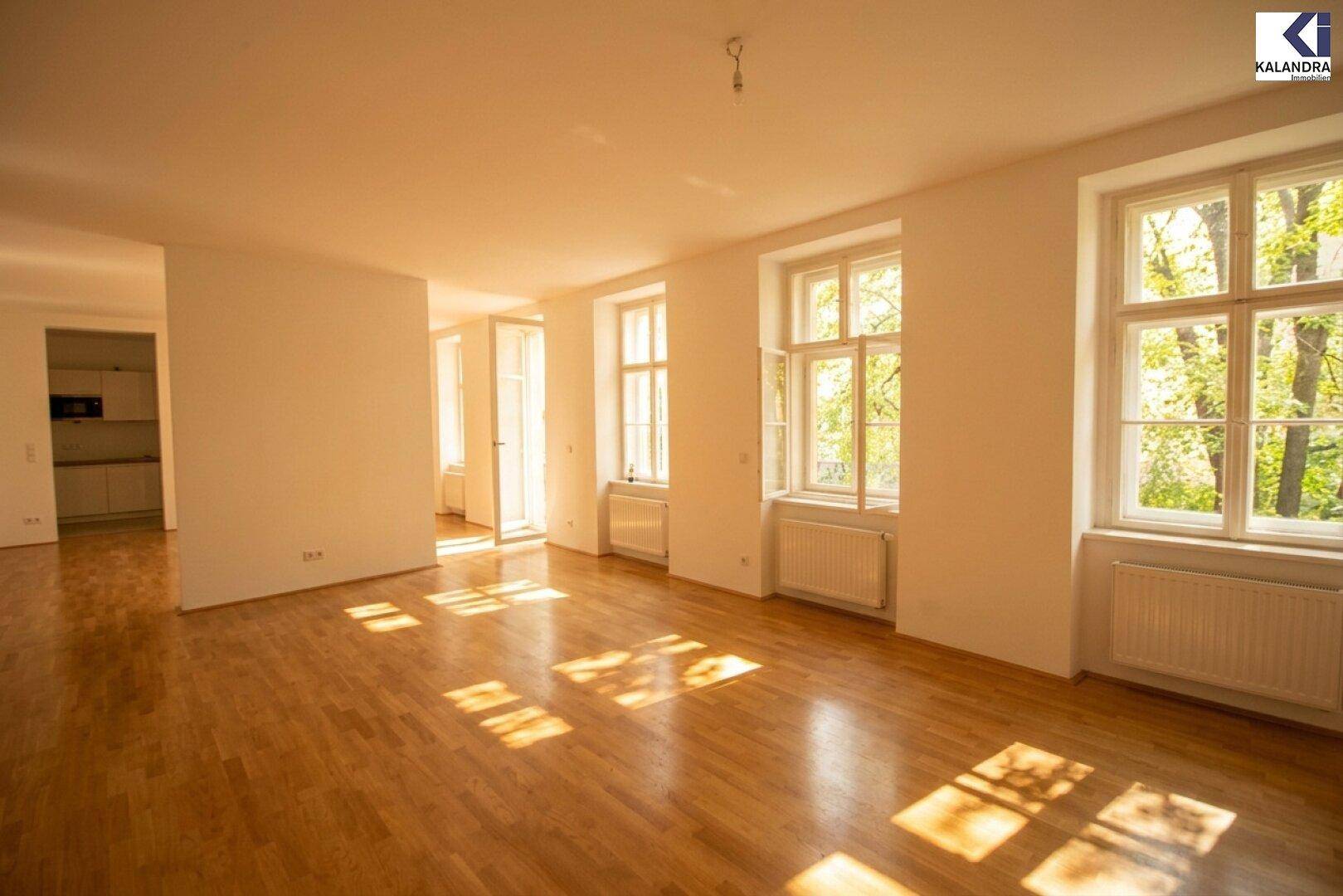 Großer Wohnbereich mit Parkettboden, vielen Fenstern und sonnigen Lichtflecken auf dem Boden.