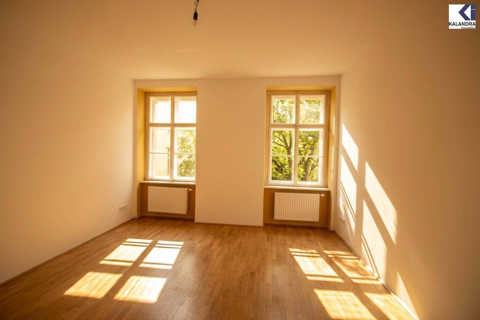 Helles Zimmer mit Parkettboden und zwei Fenstern, die den Raum mit Sonnenlicht füllen.