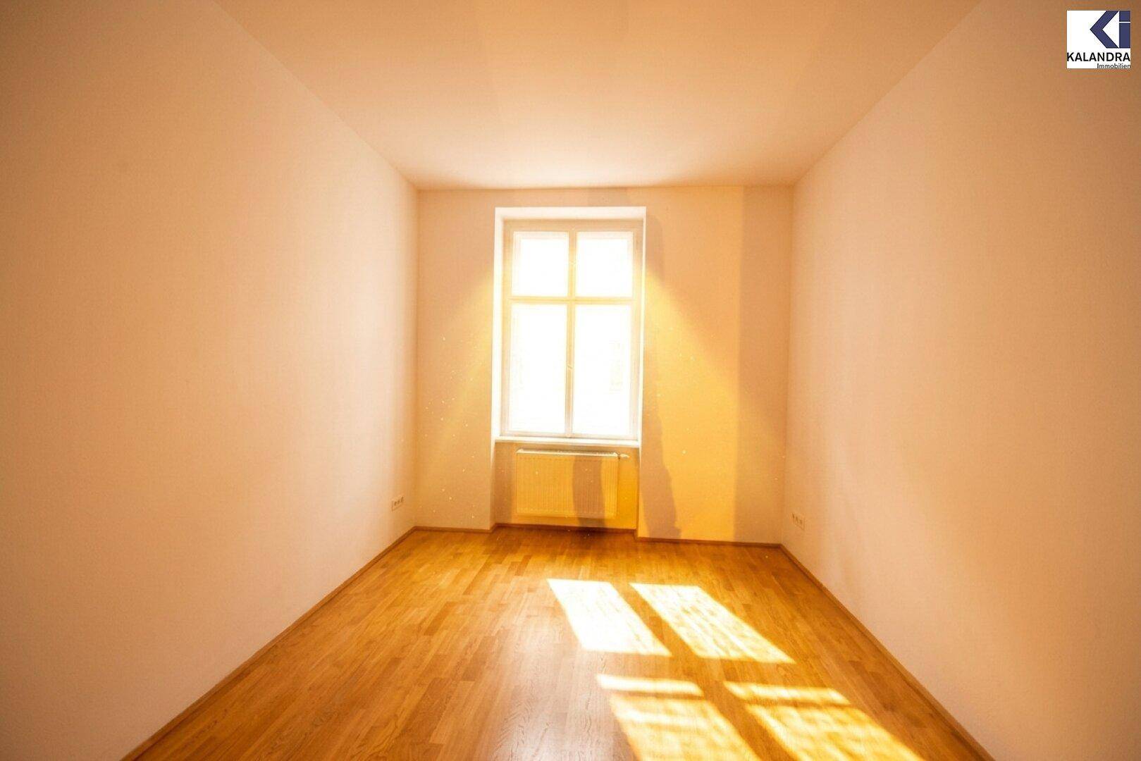Helles Zimmer mit Parkettboden und einem großen Fenster, das den Raum erhellt.
