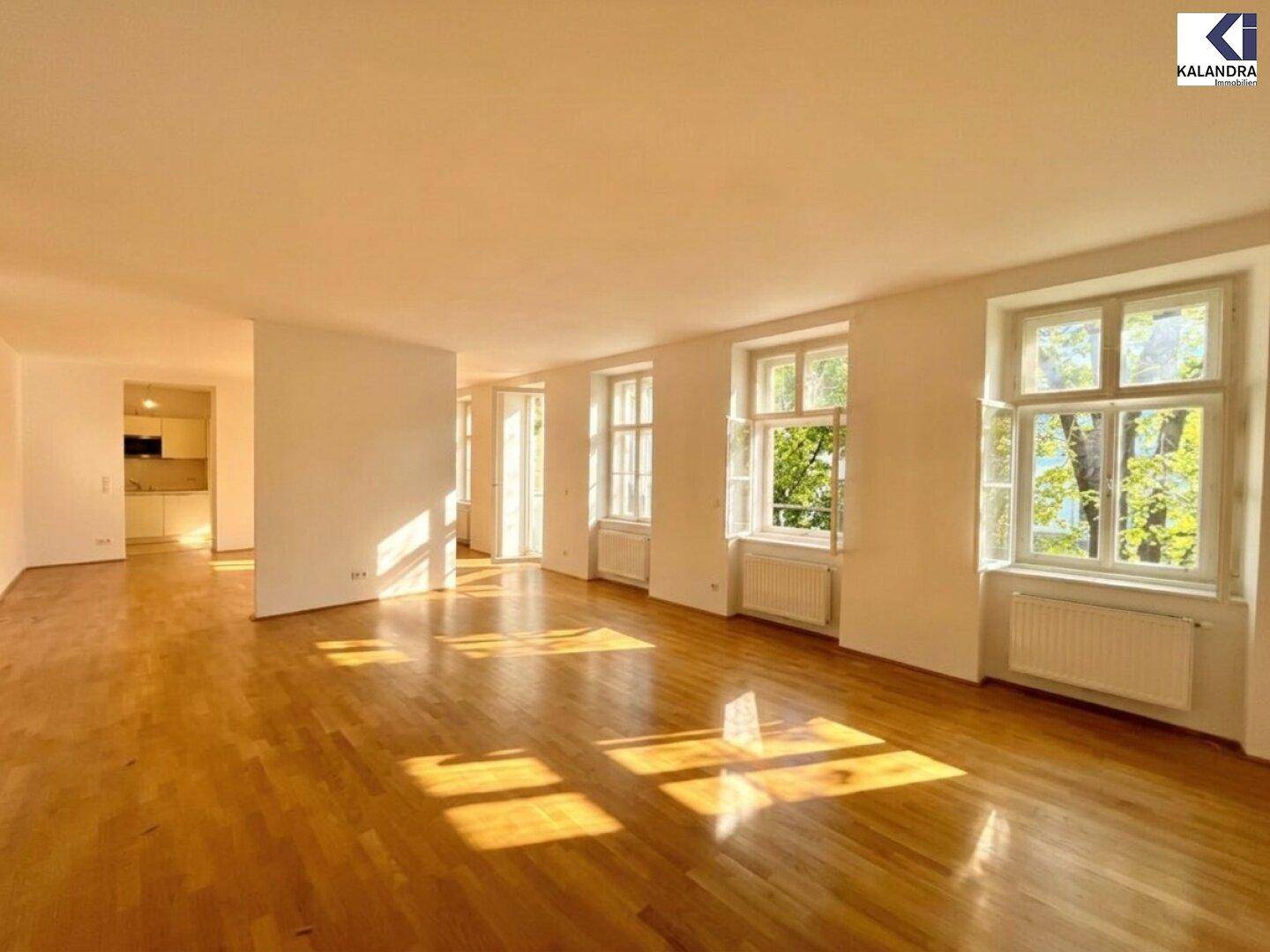 Geräumiger Wohnbereich mit Parkettboden, großen Fenstern und Blick in die angrenzende Küche.