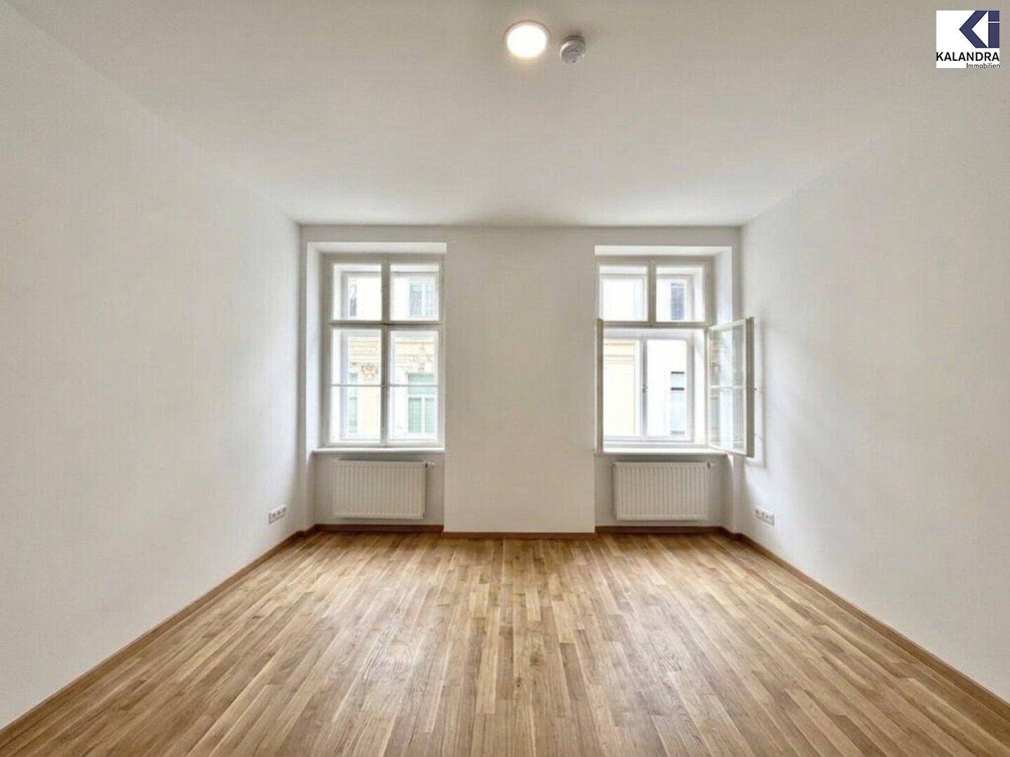 Helles Zimmer mit Parkettboden und zwei Fenstern, die den Raum mit Tageslicht versorgen.