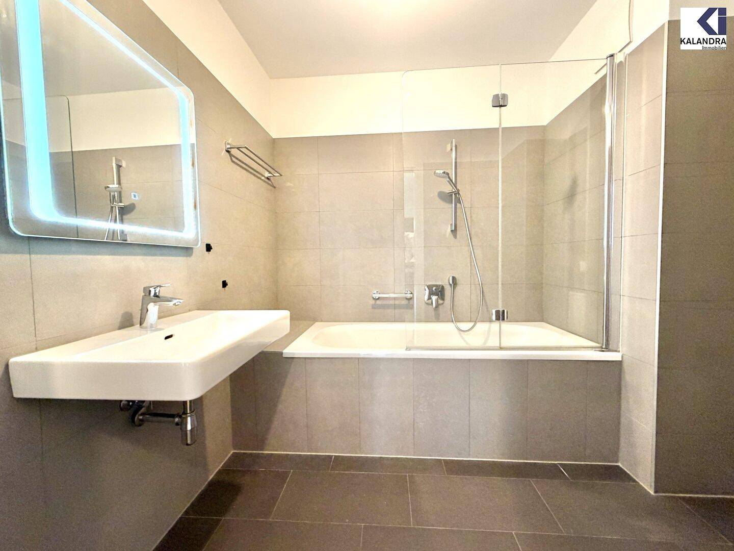 Modernes Badezimmer mit grauen Fliesen, Badewanne mit Duschwand und beleuchtetem Spiegel.