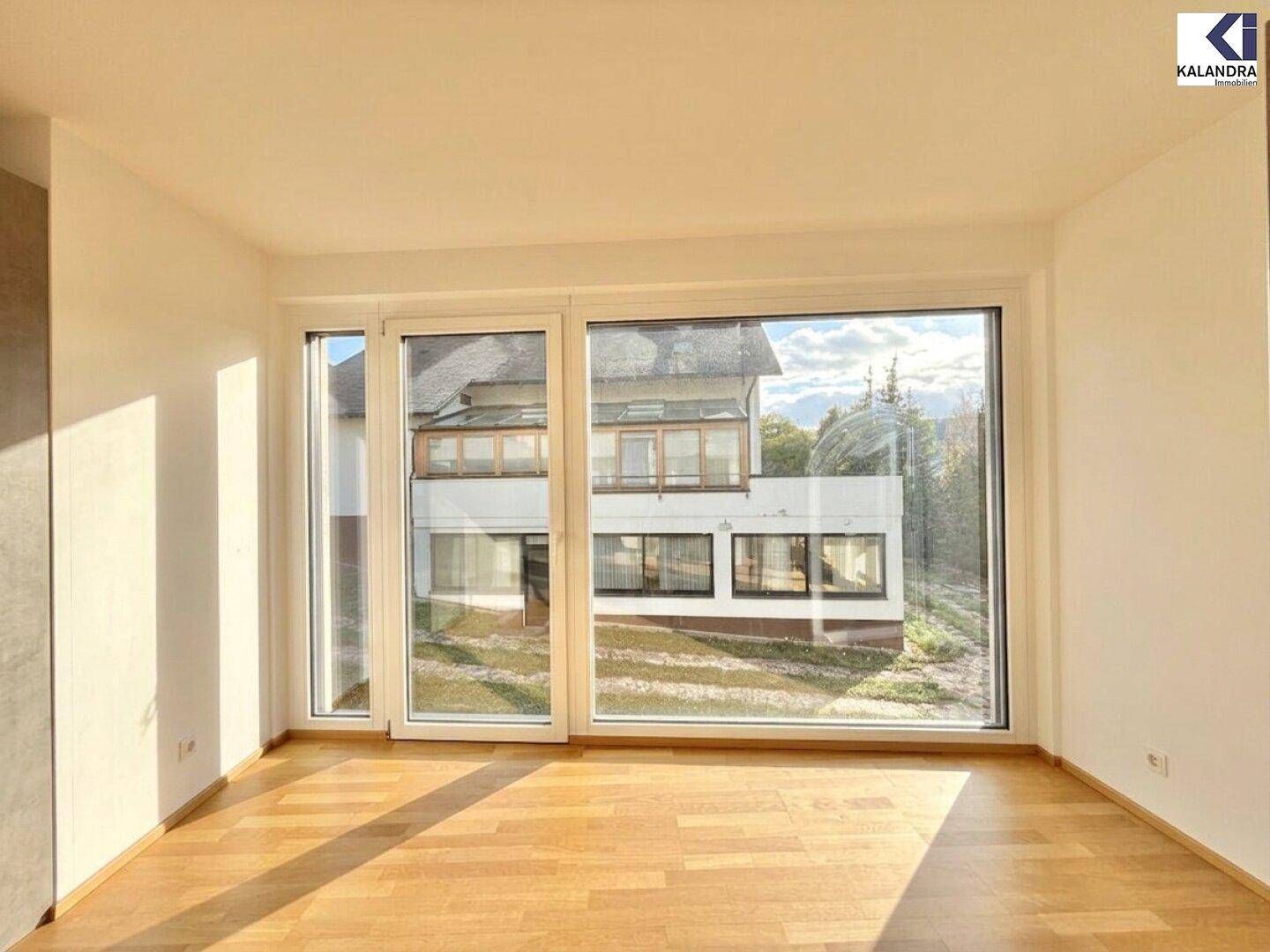 Lichtdurchfluteter Raum mit Holzboden und großen Fenstern, die einen Blick auf das Gebäude gegenüber bieten.