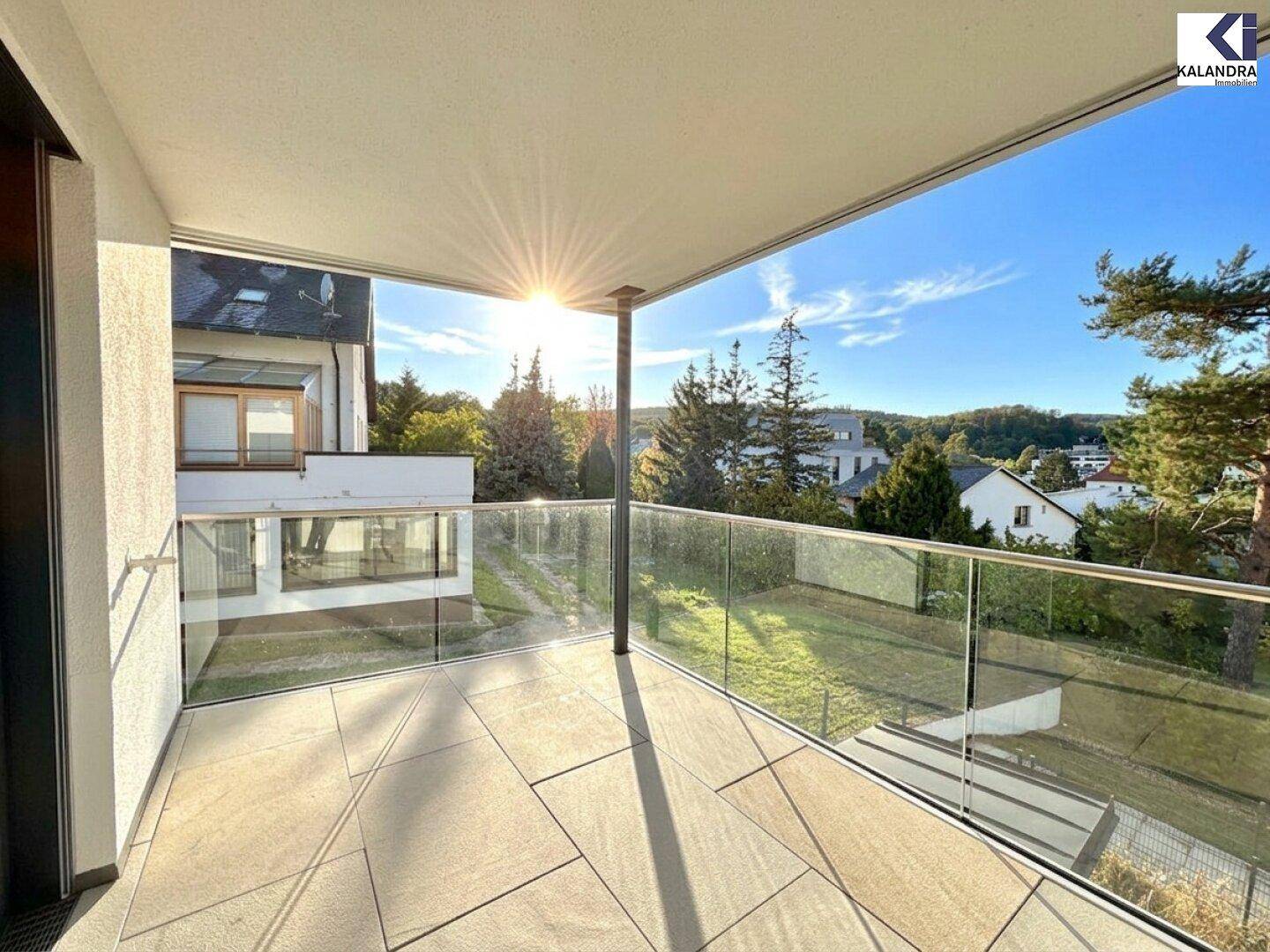 Sonniger Balkon mit Glasgeländer und Blick auf die grüne Umgebung und andere Häuser.