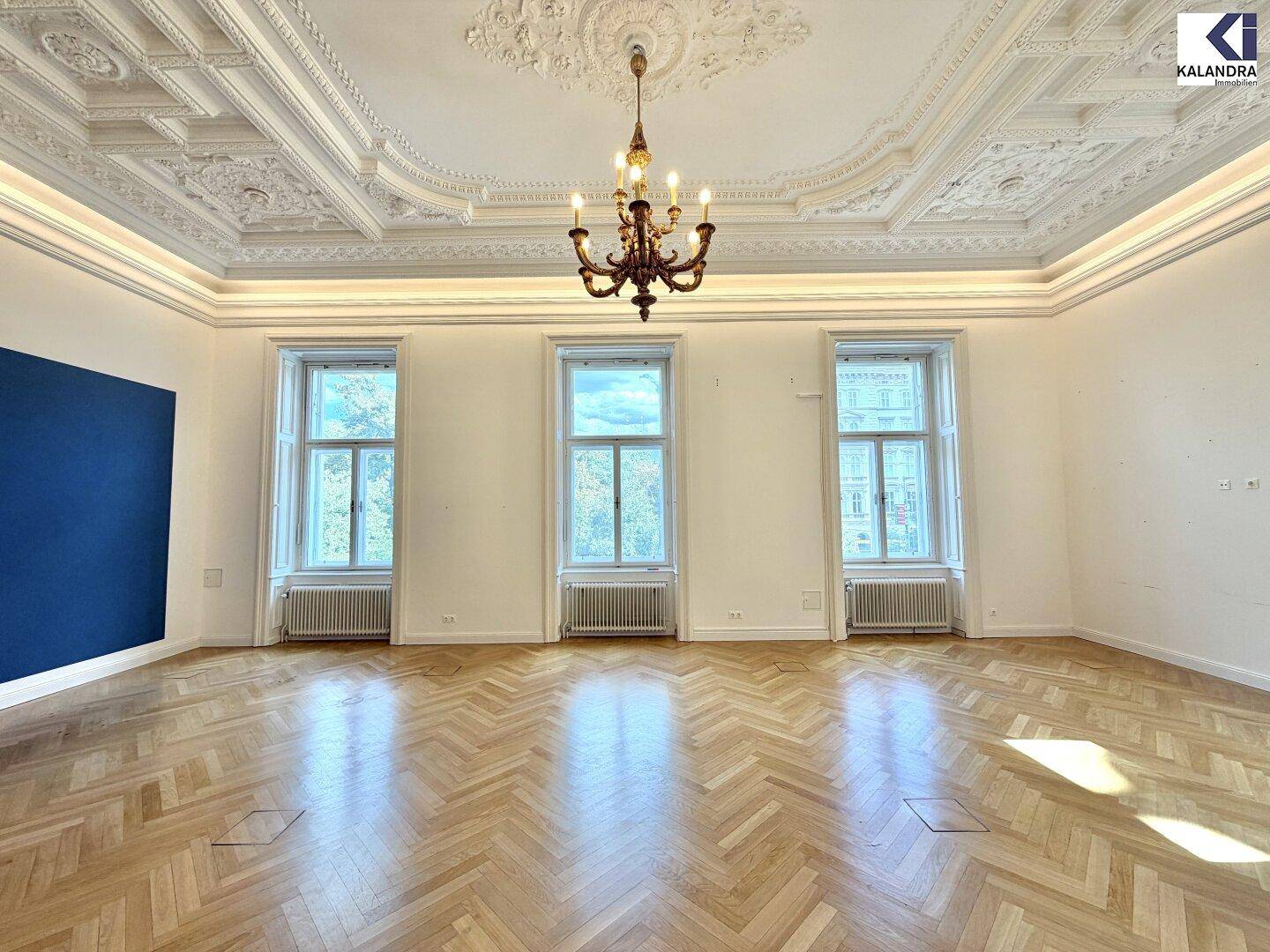 Heller Raum mit Fischgrätparkett, hohen Decken und Stuckverzierungen, ergänzt durch einen klassischen Kronleuchter.